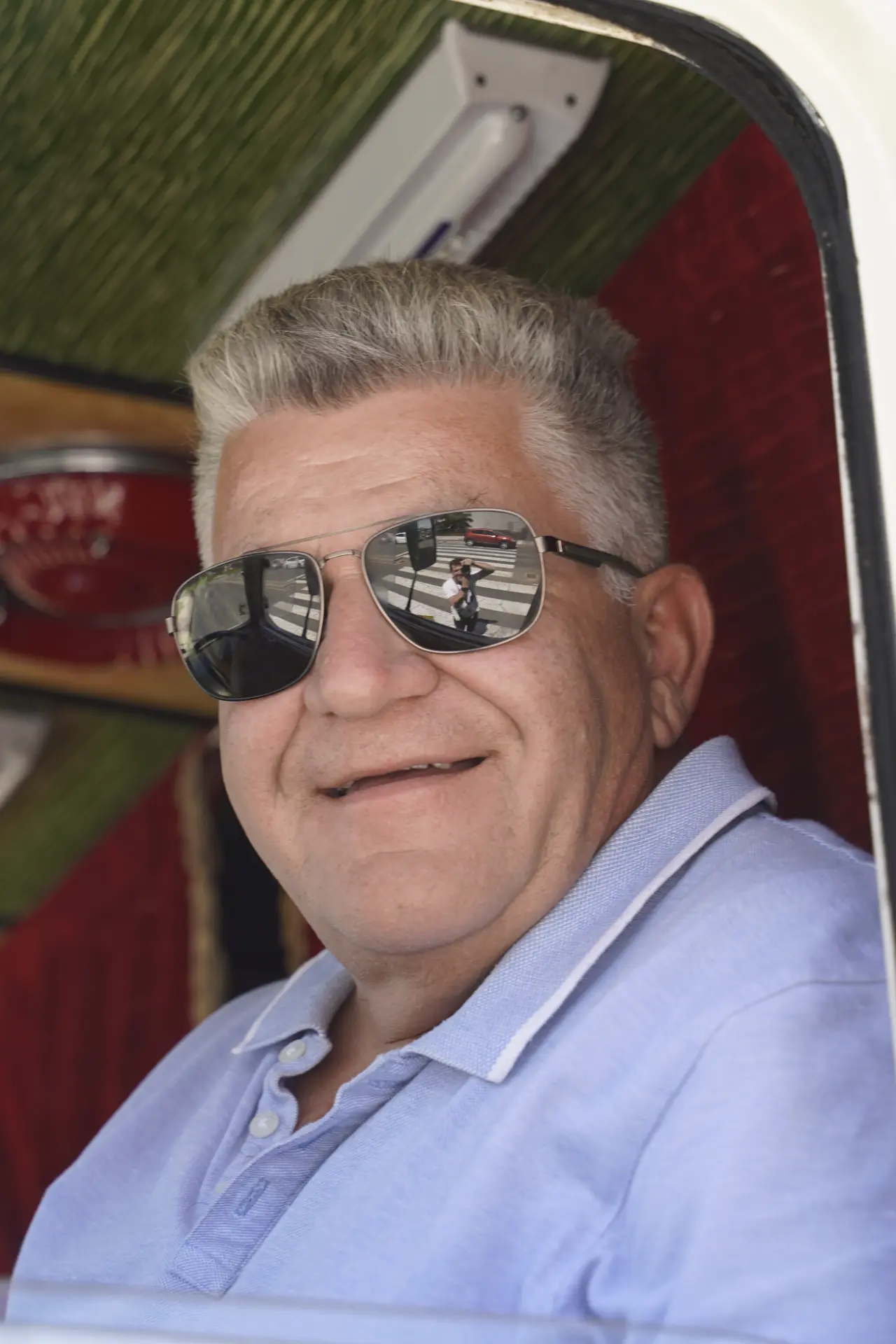 Hombre sonriente con gafas de sol y camisa azul claro sentado en el interior de un vehículo en la concentración solidaria de camiones; reflejo de otros vehículos visible en sus gafas.