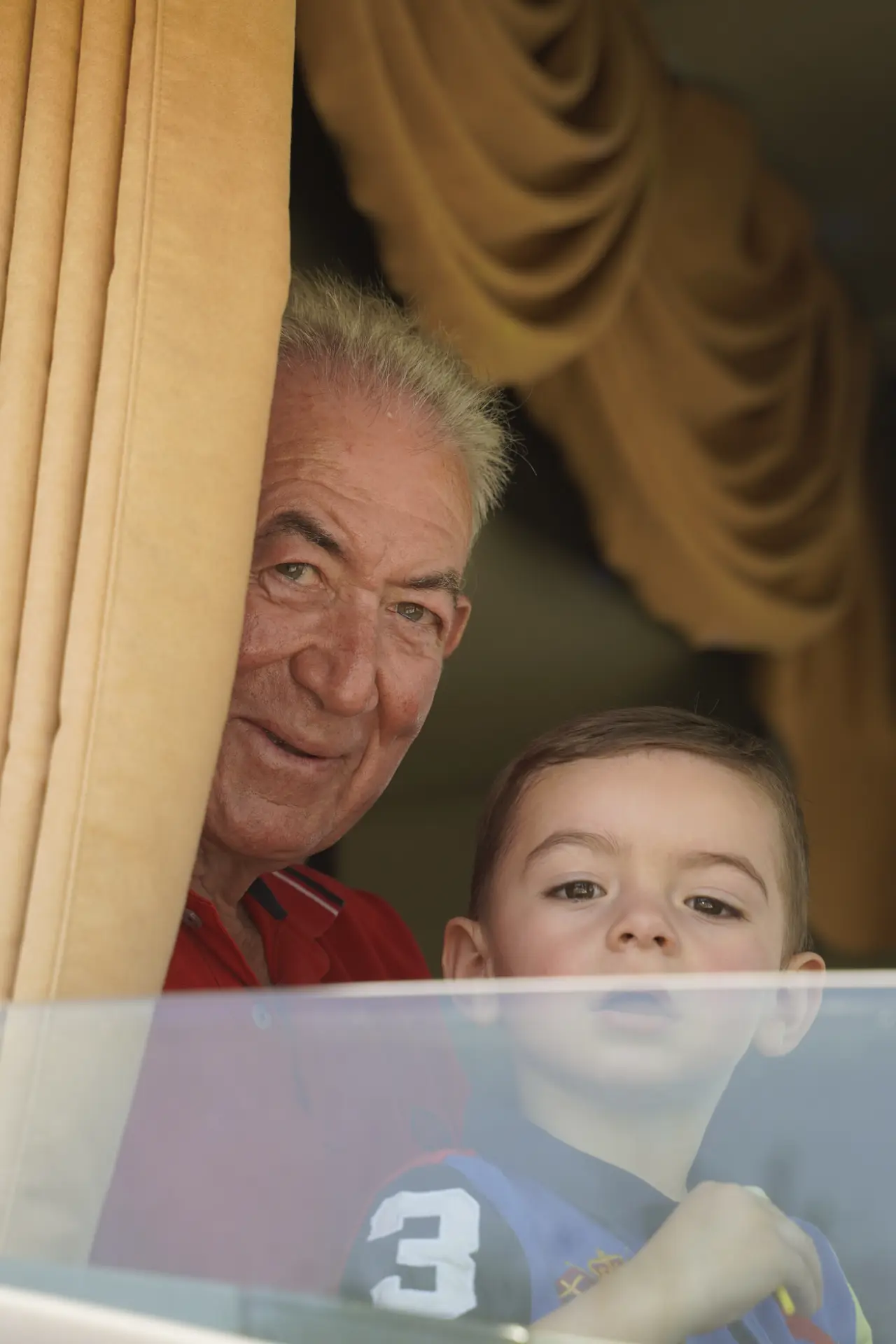 Un hombre mayor y un niño miran desde detrás de unas cortinas doradas, ambos sonríen amablemente hacia la cámara en una concentración solidaria de camiones en Ávila.