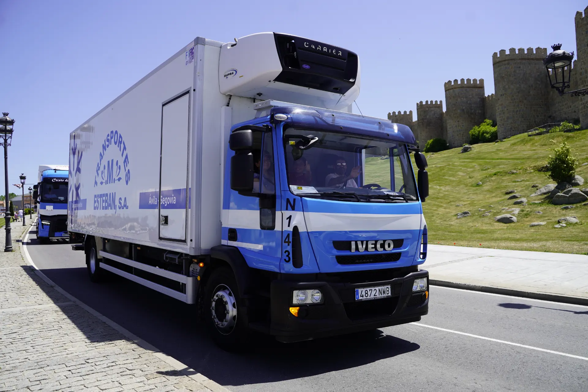 Un camión frigorífico Iveco azul y blanco, perteneciente a la concentración solidaria de camiones, circula en un día soleado cerca de la muralla del histórico castillo de piedra de Ávila.