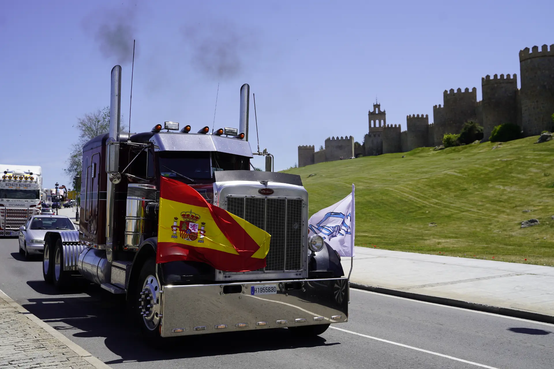 Un semirremolque con banderas españolas pasa por delante de la muralla del histórico castillo de Ávila en un día soleado, capturando el espíritu de una concentración solidaria de camiones.
