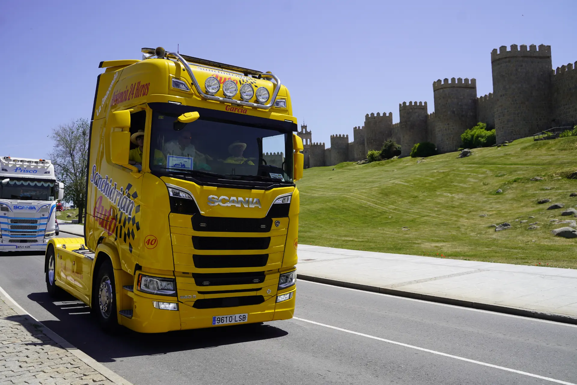 Un camión Scania amarillo brillante circula por una carretera junto a un histórico muro de piedra y una verde colina cubierta de hierba en Ávila, uniéndose a otros camiones para una concentración solidaria de camiones.