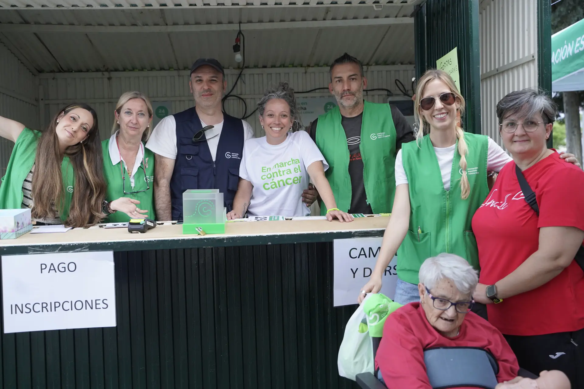 Ocho voluntarios sonrientes con chalecos verdes en un puesto al aire libre de concienciación sobre el cáncer e inscripción en un evento durante una concentración solidaria de camiones en Ávila, con varios vehículos visibles al fondo.
