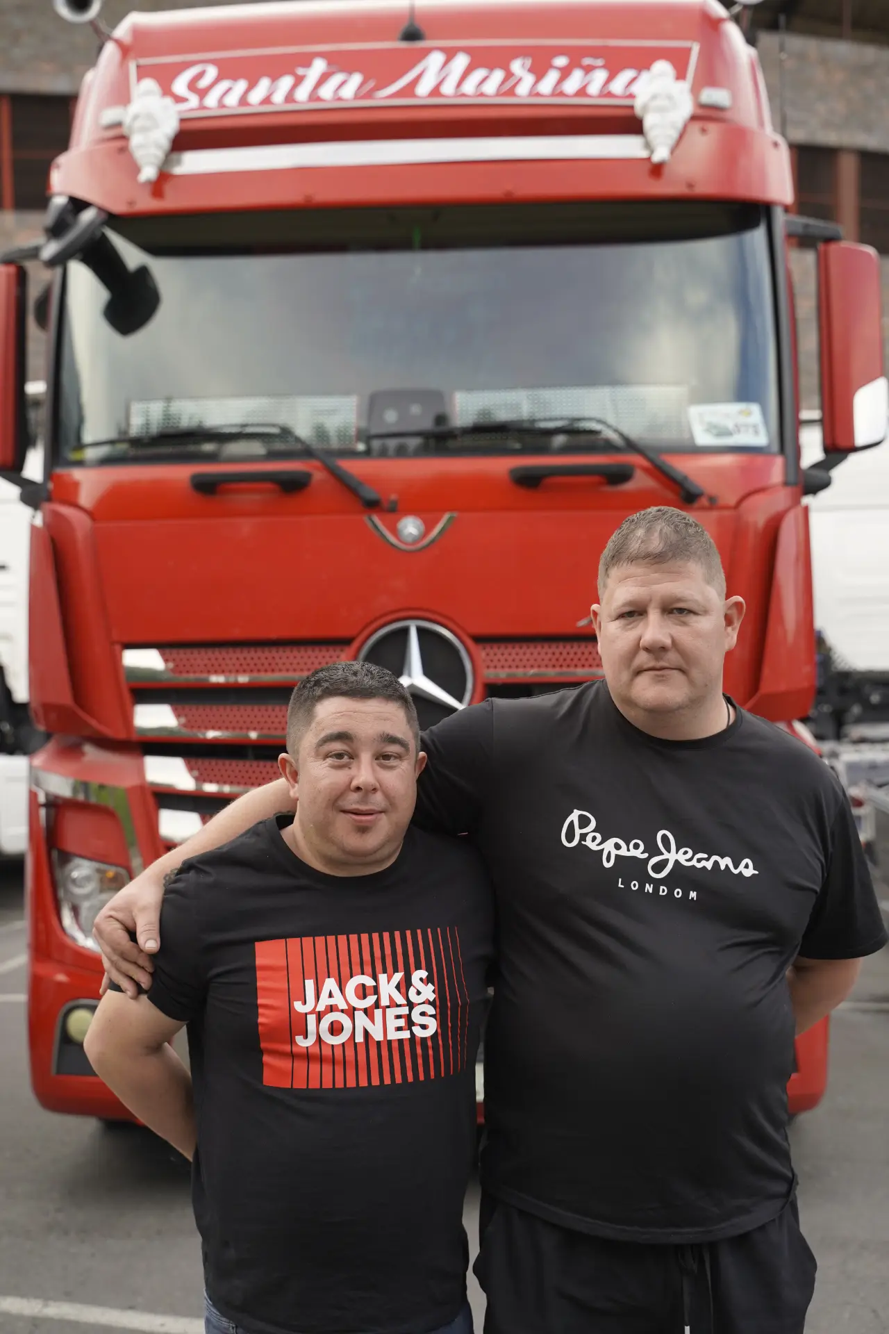 Dos hombres del brazo delante de un camión rojo con la inscripción "Santa Marina" en la parte superior, durante una concentración solidaria de camiones en Ávila, rodeados de otros vehículos.
