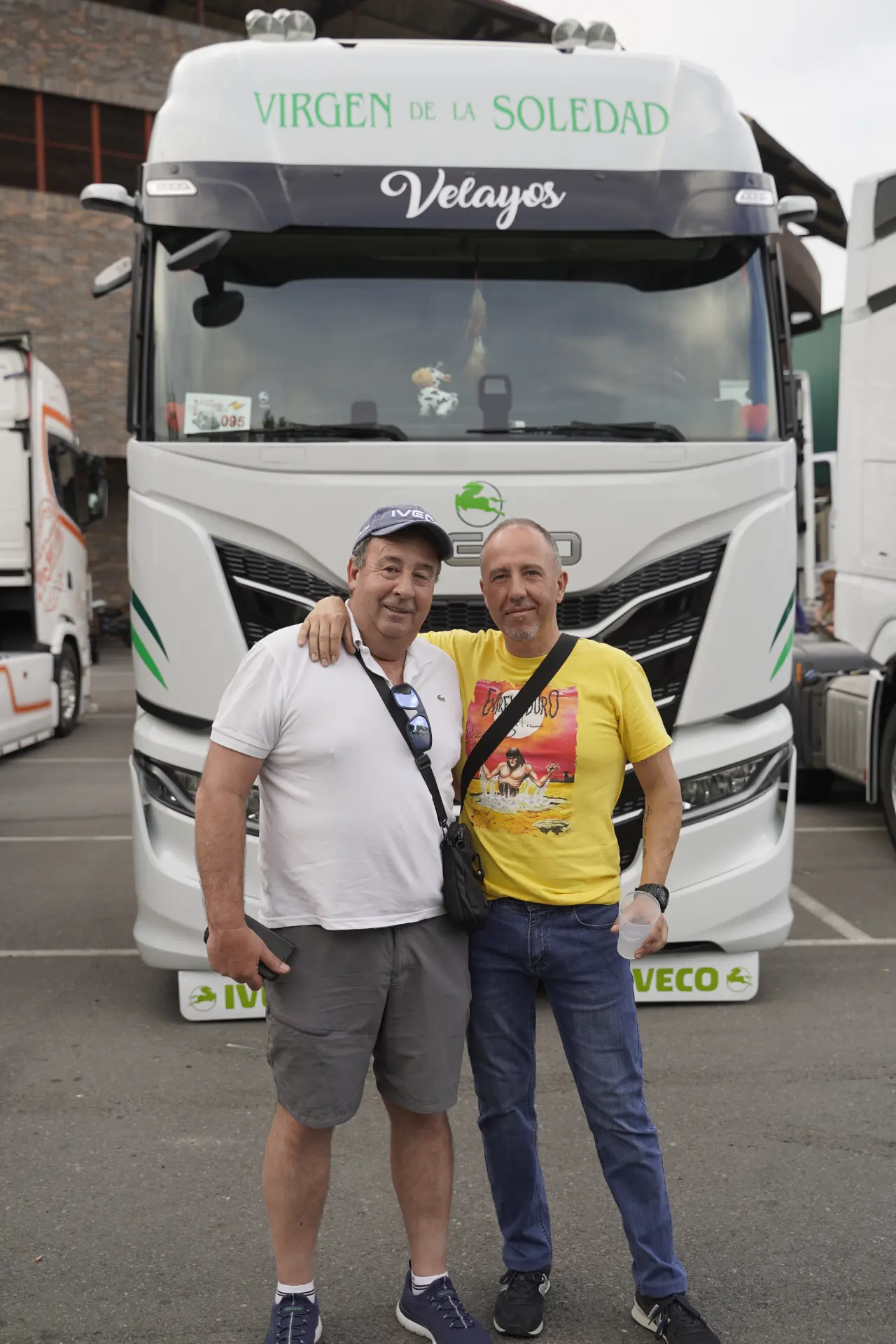 Dos hombres posan sonrientes delante de un gran camión blanco con la inscripción "Virgen de la Soledad" en una concentración solidaria de camiones en Ávila.