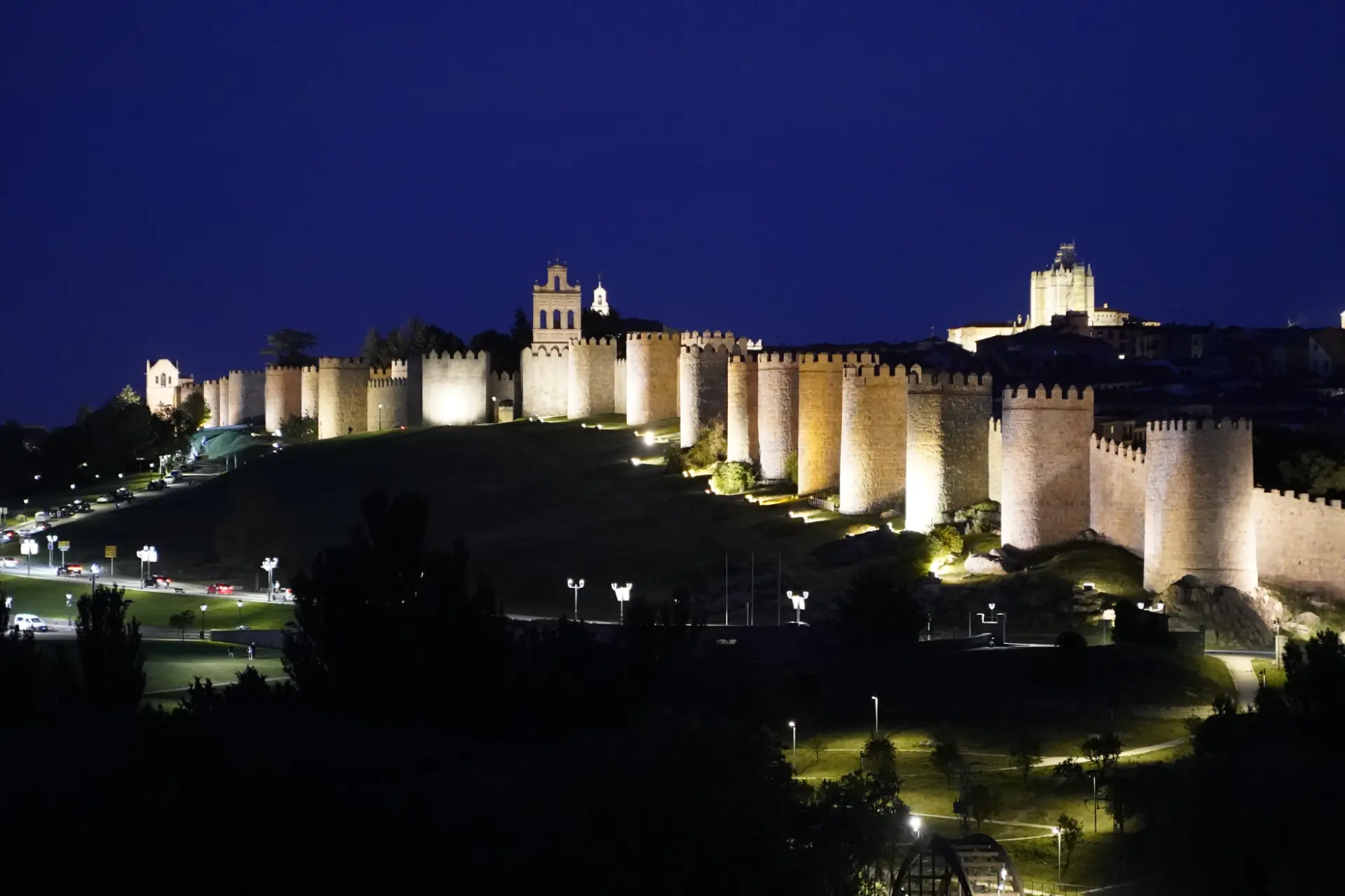 Las antiguas murallas de piedra de Ávila se iluminan por la noche, con torres y edificios visibles al fondo, mientras los vehículos se reúnen en las inmediaciones para una concentración solidaria de camiones.
