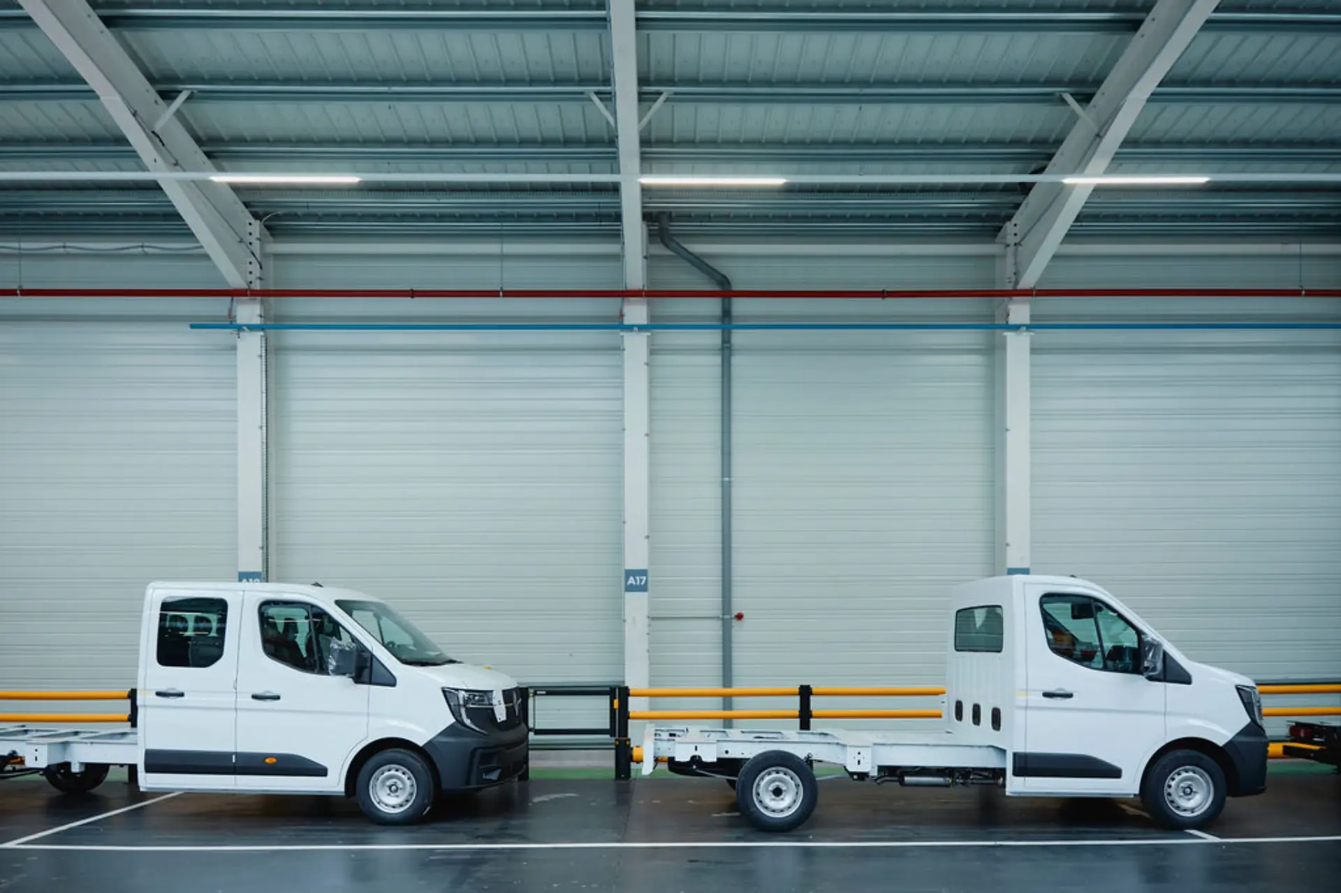 Dos furgonetas blancas con la carrocería sin terminar, a la espera de ser personalizadas, están aparcadas una al lado de la otra en un garaje industrial cubierto.