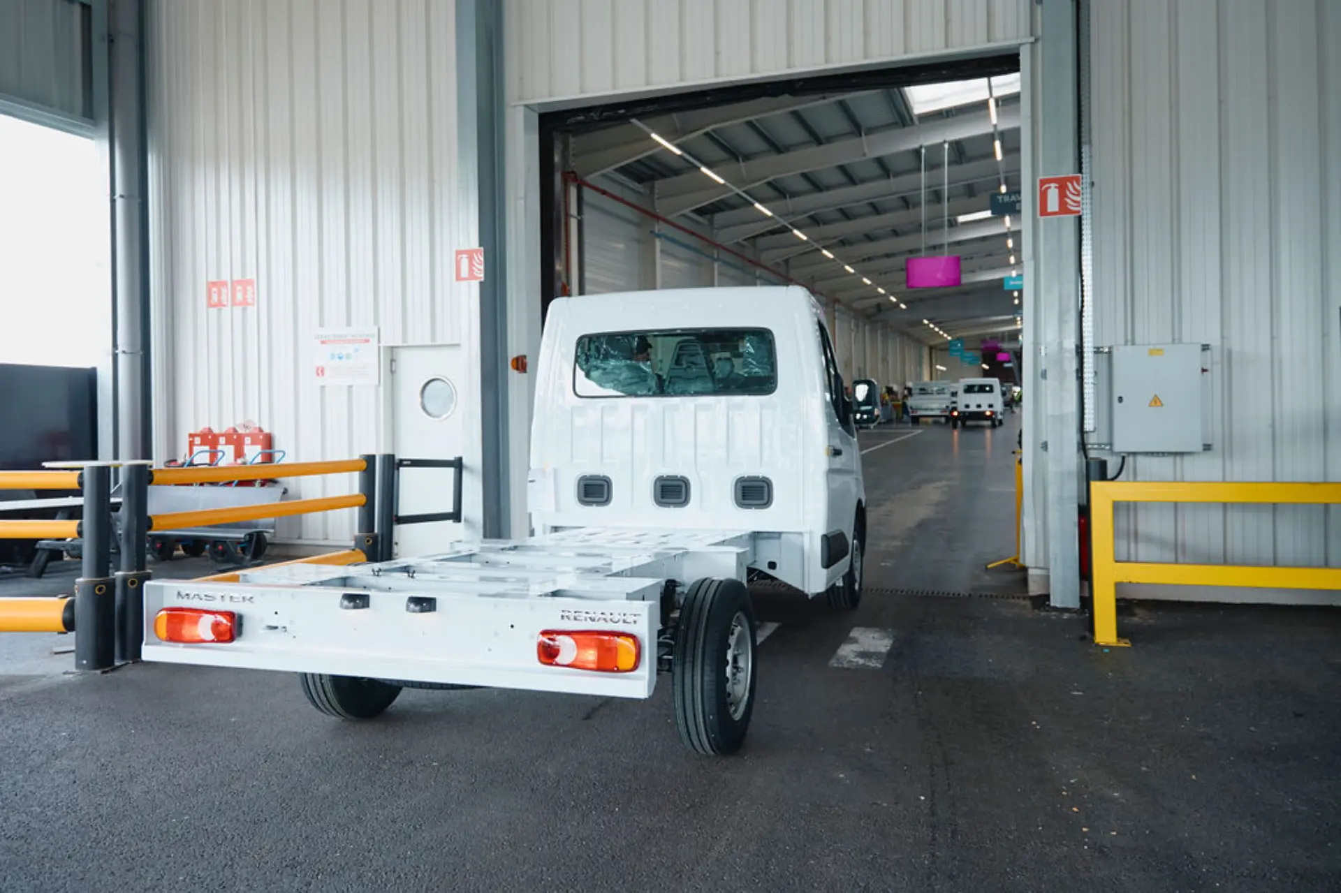 Un camión de plataforma blanca de la marca Renault, transformado por la propia Renault, está entrando marcha atrás en un almacén repleto de otros vehículos industriales.