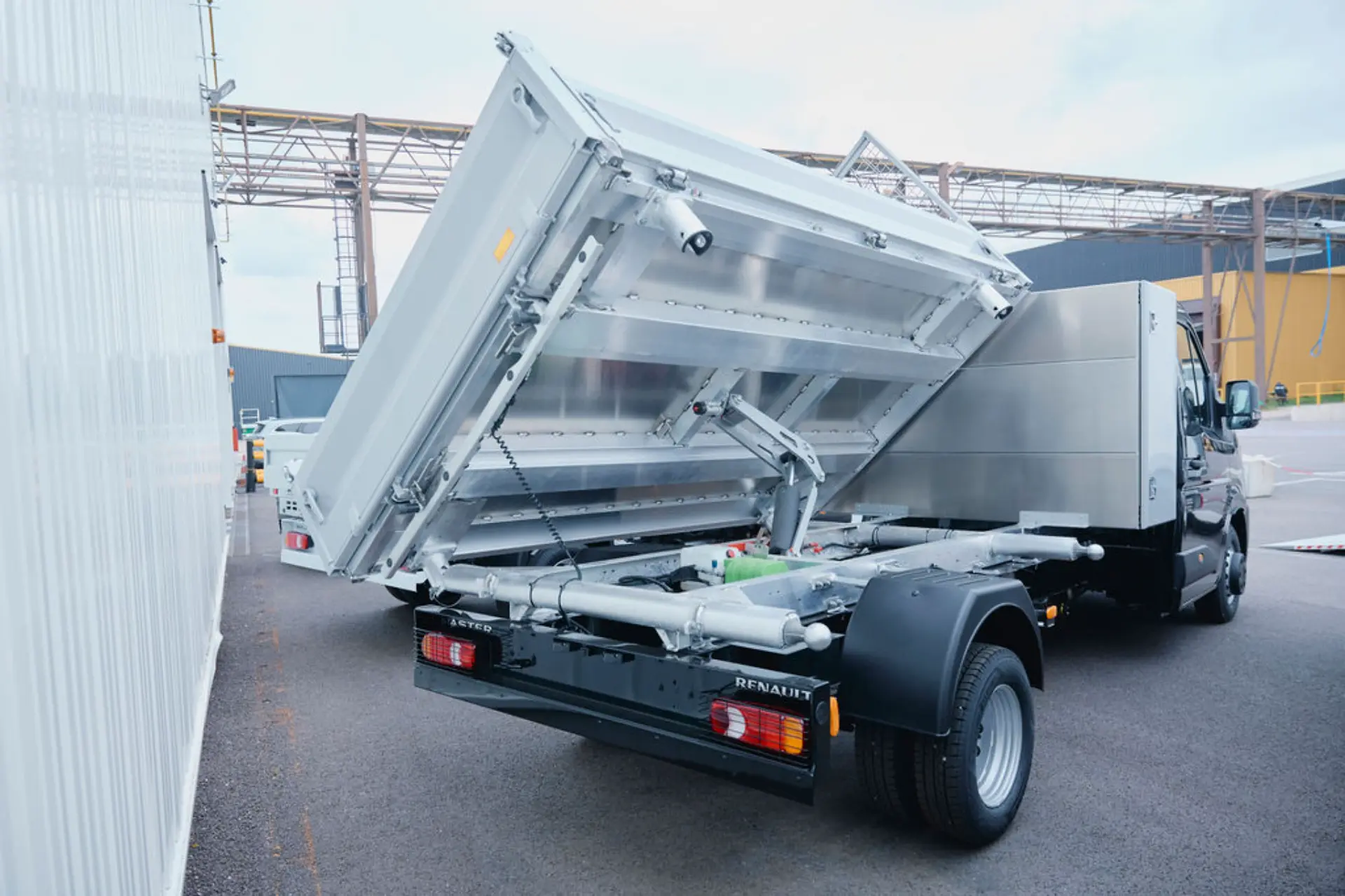 Un camión volquete plateado, un claro ejemplo de vehículos industriales, con la caja elevada en un recinto industrial, muestra su mecanismo de elevación hidráulico.