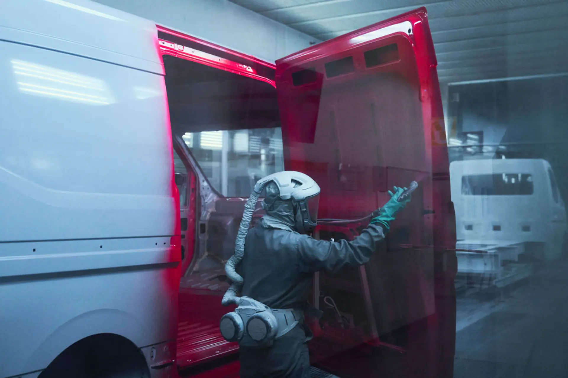 Una persona con equipo de protección pintando con spray las puertas traseras abiertas de una furgoneta Renault Master blanca en el interior de un taller en Batilly.