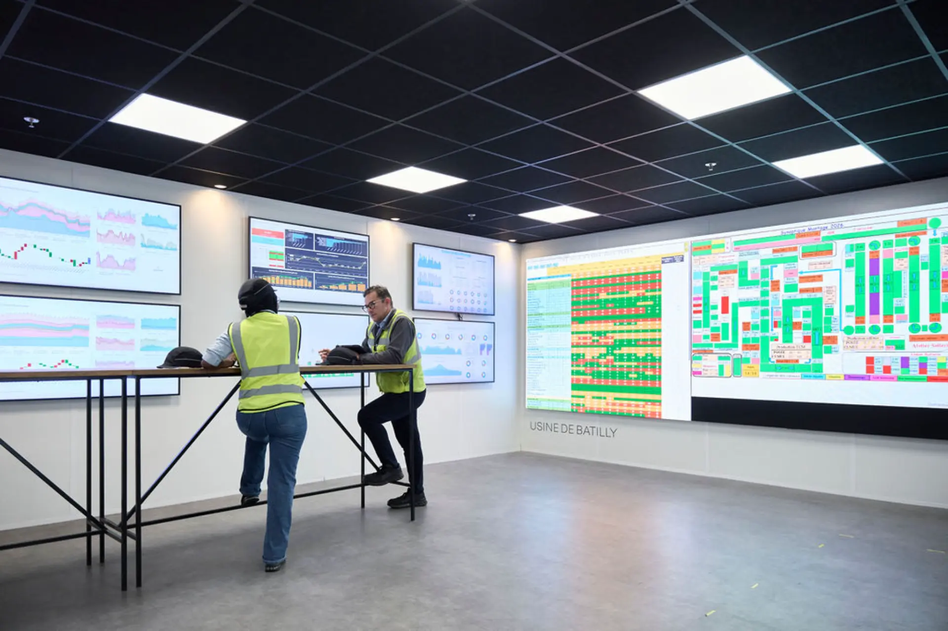 Dos personas con chalecos de seguridad revisan datos en sus ordenadores portátiles en una moderna sala de control dotada de grandes pantallas digitales, supervisando las operaciones de la producción del Renault Master en Batilly.