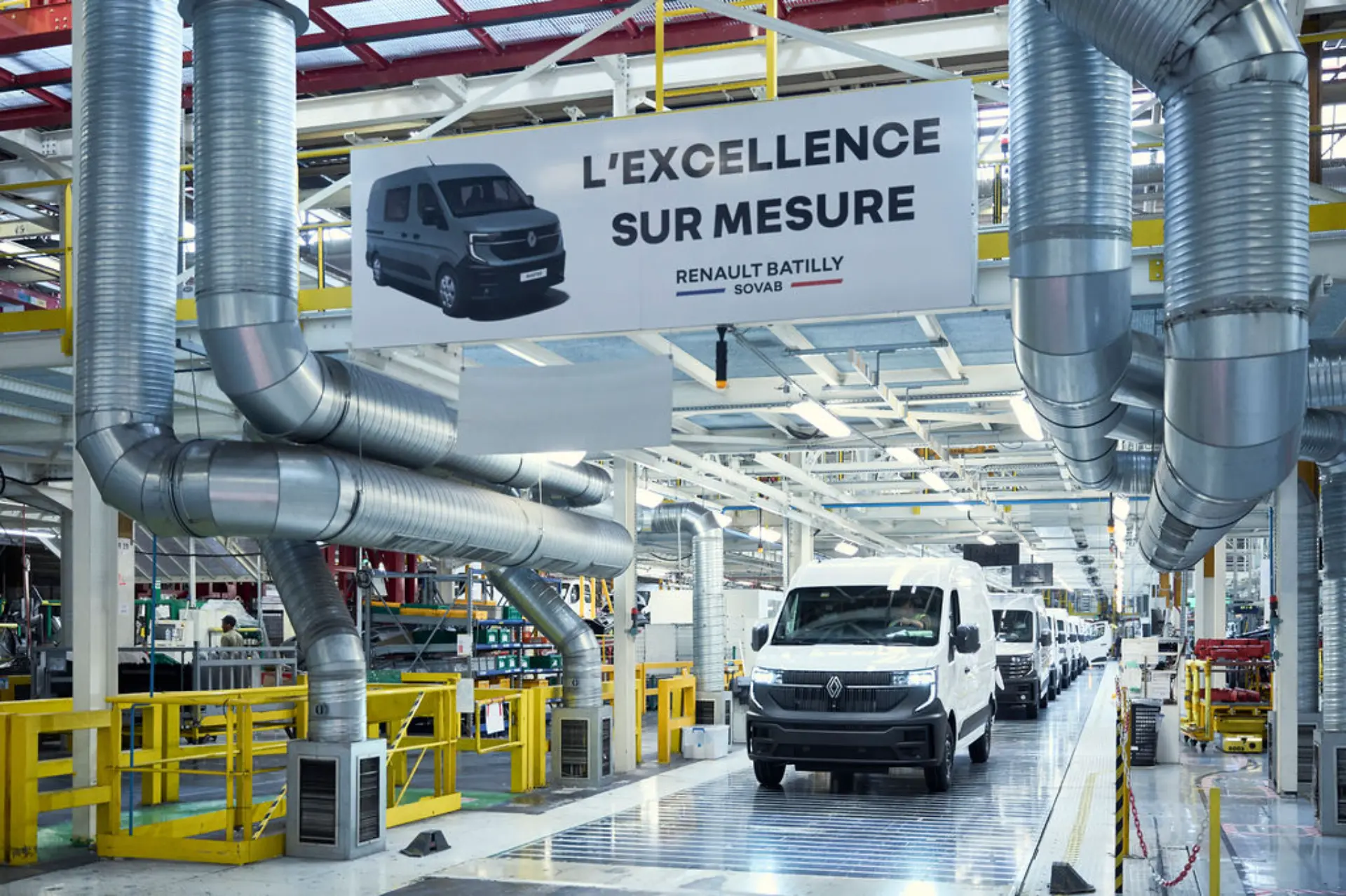 Furgonetas Renault blancas en una cadena de montaje de una fábrica, con un cartel que reza «L'excellence sur mesure», que destaca la personalización que ofrece Renault para cada vehículo.