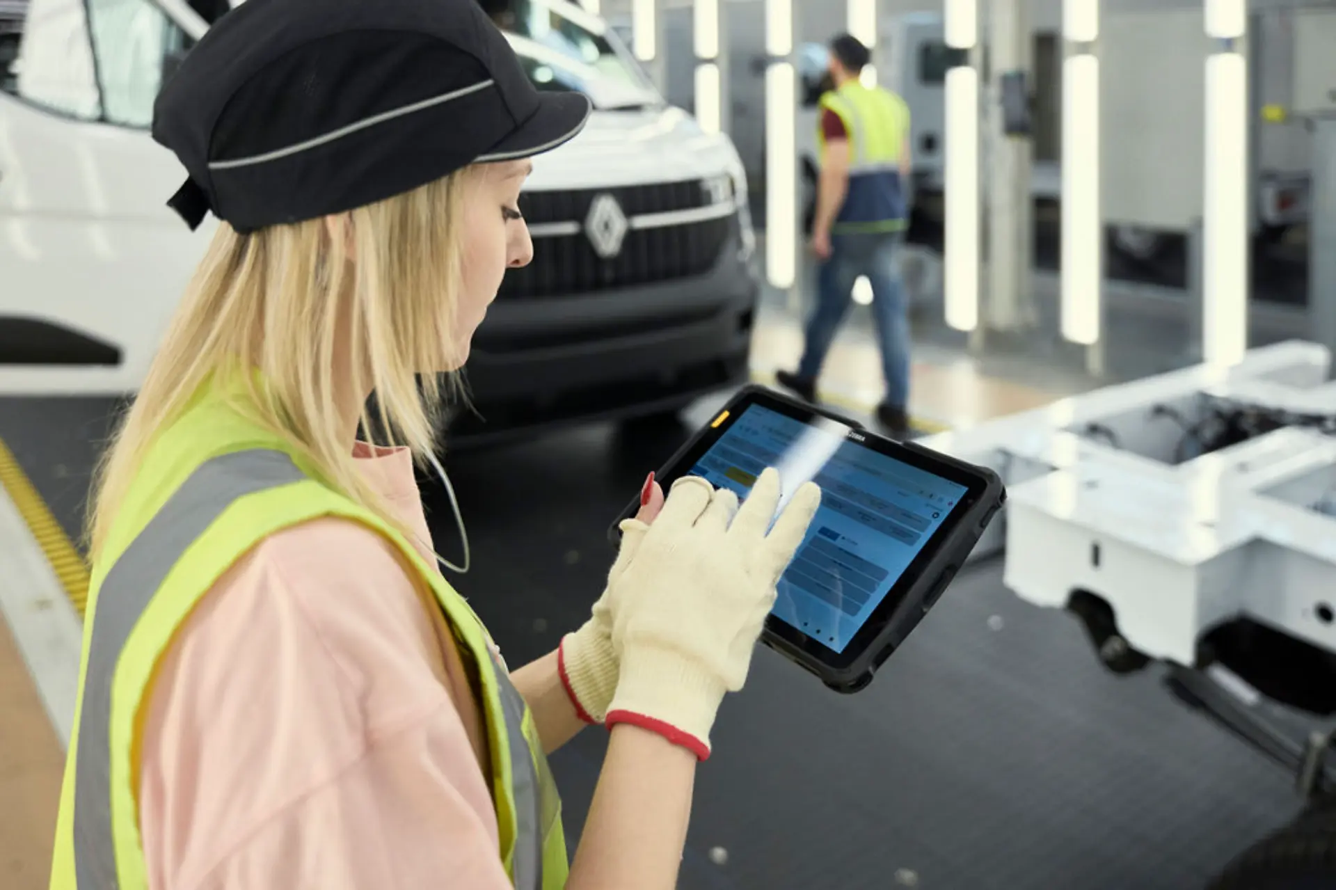 Una mujer con equipo de seguridad utiliza una tableta en la cadena de montaje del Renault Master en la planta de Batilly, con coches y trabajadores al fondo.