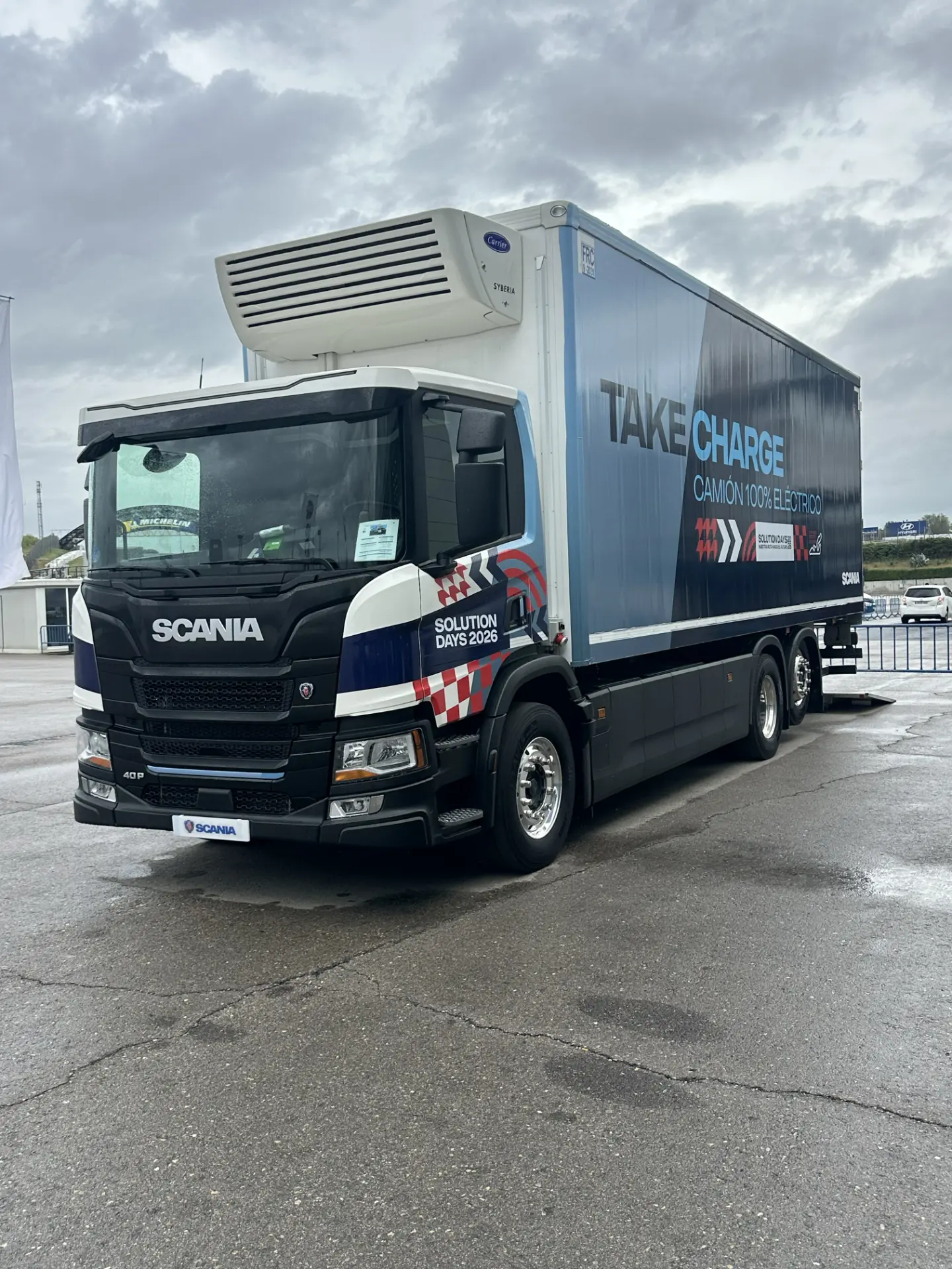 Un camión eléctrico Scania con los lemas «Take Charge» y «100 % eléctrico» está aparcado sobre el asfalto mojado, lo que pone de manifiesto cómo los camiones Scania marcan el camino en soluciones de transporte innovadoras.