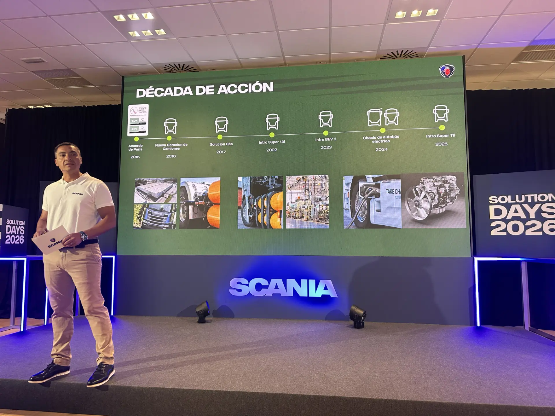Un hombre se encuentra en el escenario junto a una gran pantalla en la que se muestra la cronología de la «Década de Acción» de Scania durante el evento Scania Solution Days.