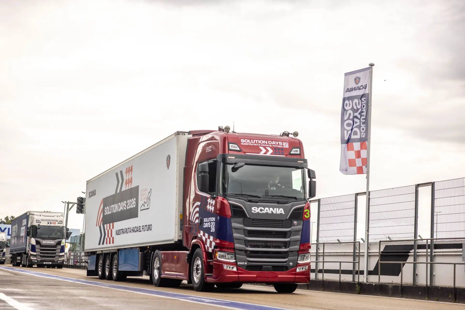 Un camión Scania rojo y blanco circula por una carretera junto a la valla y la bandera de un circuito de carreras, mostrando las robustas soluciones de Scania para cualquier reto de transporte.
