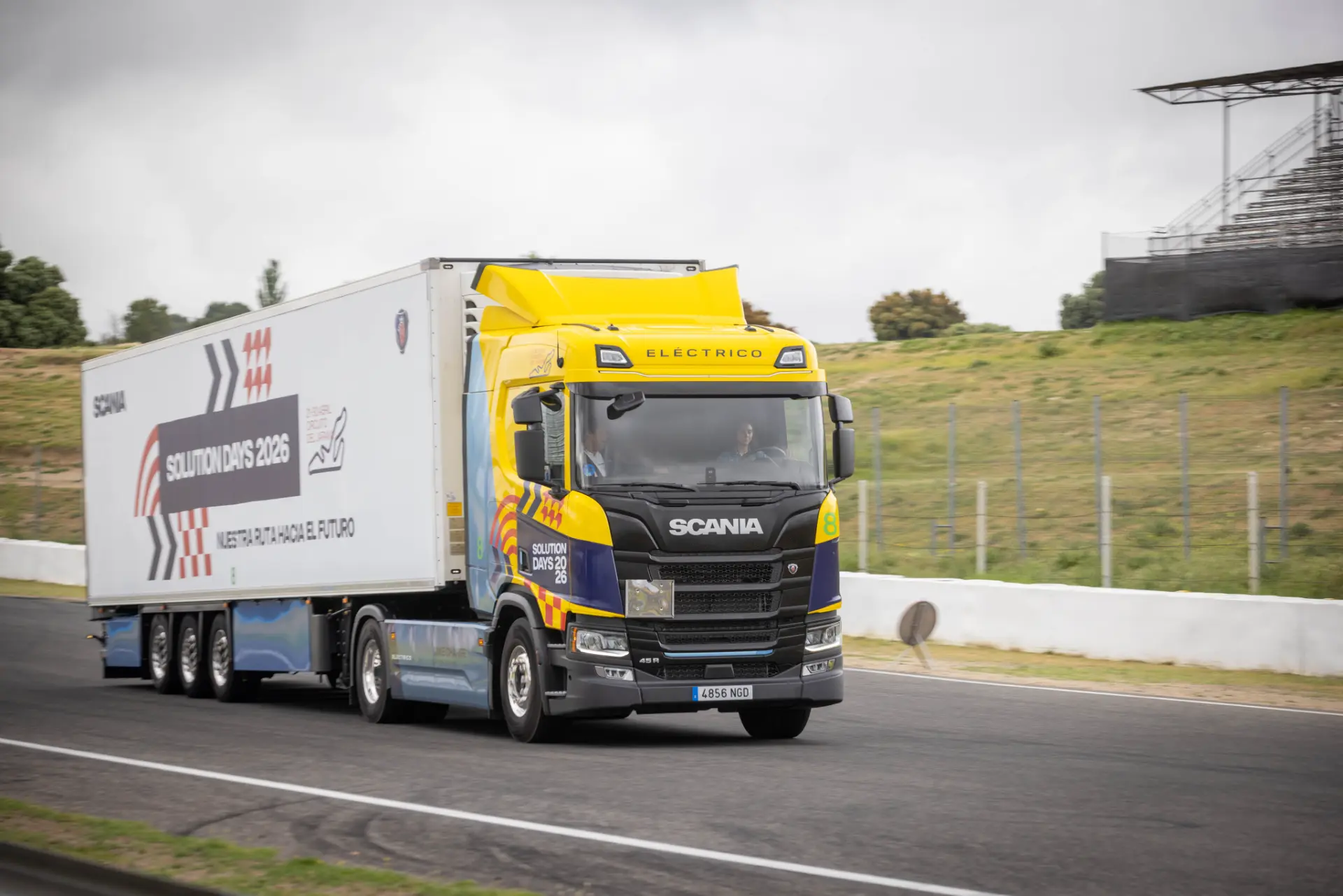 Un camión eléctrico Scania de color amarillo y azul circula por un circuito con un remolque blanco, mostrando la innovación de los camiones Scania.