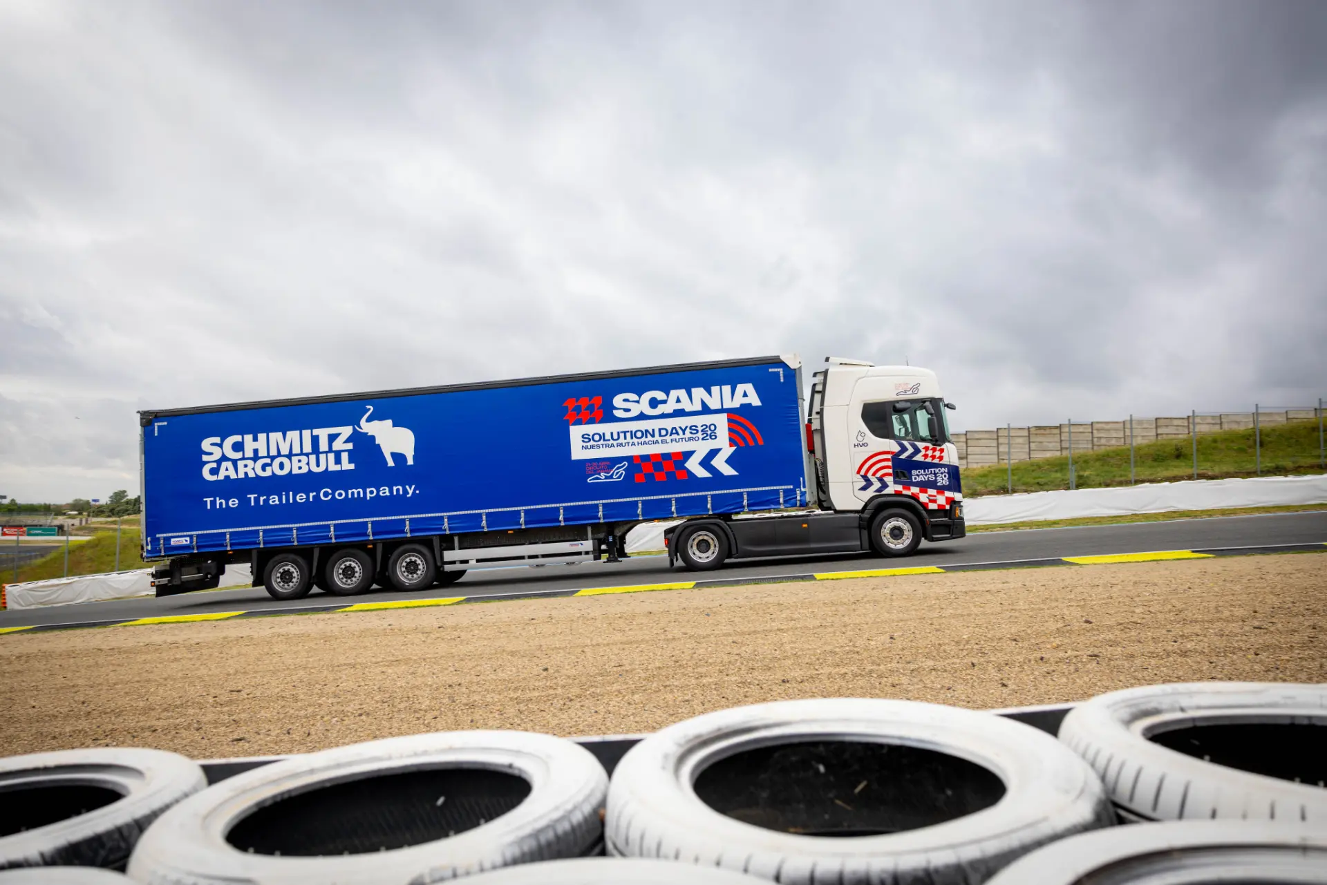 Un camión azul de Schmitz Cargobull y Scania circula por una pista junto a barreras de neumáticos bajo un cielo nublado durante los Scania Solution Days, en los que se presentan las innovadoras soluciones de Scania para camiones Scania.