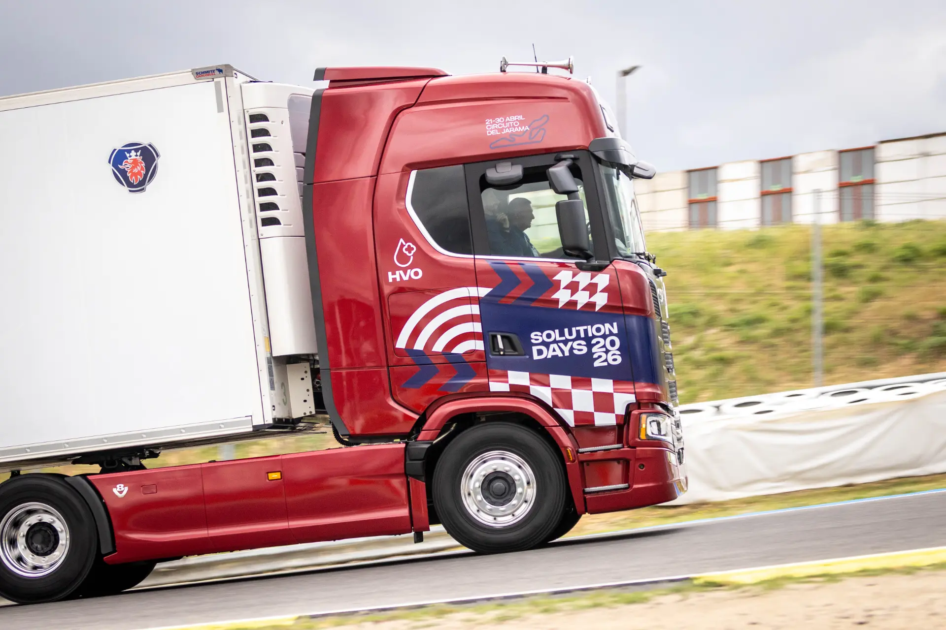 Un camión Scania rojo con el letrero «Solution Days 2026» en el lateral circula por una carretera junto a una zona verde, lo que pone de manifiesto la fiabilidad de los camiones Scania.