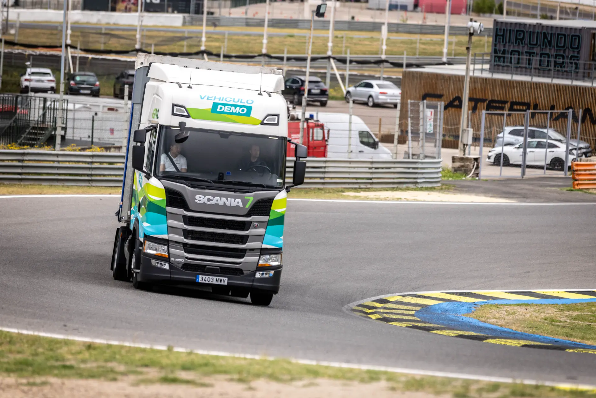 Un camión de demostración de Scania recorre un circuito de carreras con barreras y coches aparcados al fondo durante los Scania Solution Days, en los que se presentan las innovaciones de los camiones Scania.