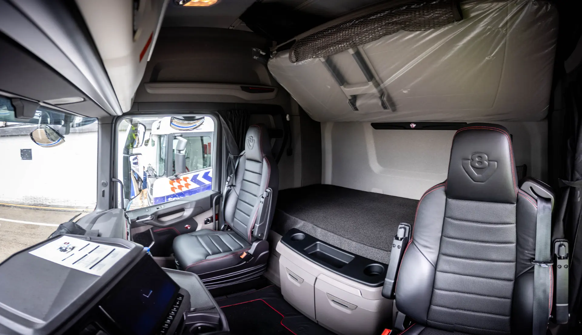 Interior de una moderna cabina de camión Scania con dos asientos negros y una cama situada detrás del puesto del conductor, que destaca por su comodidad e innovación inspiradas en Soluciones Scania.