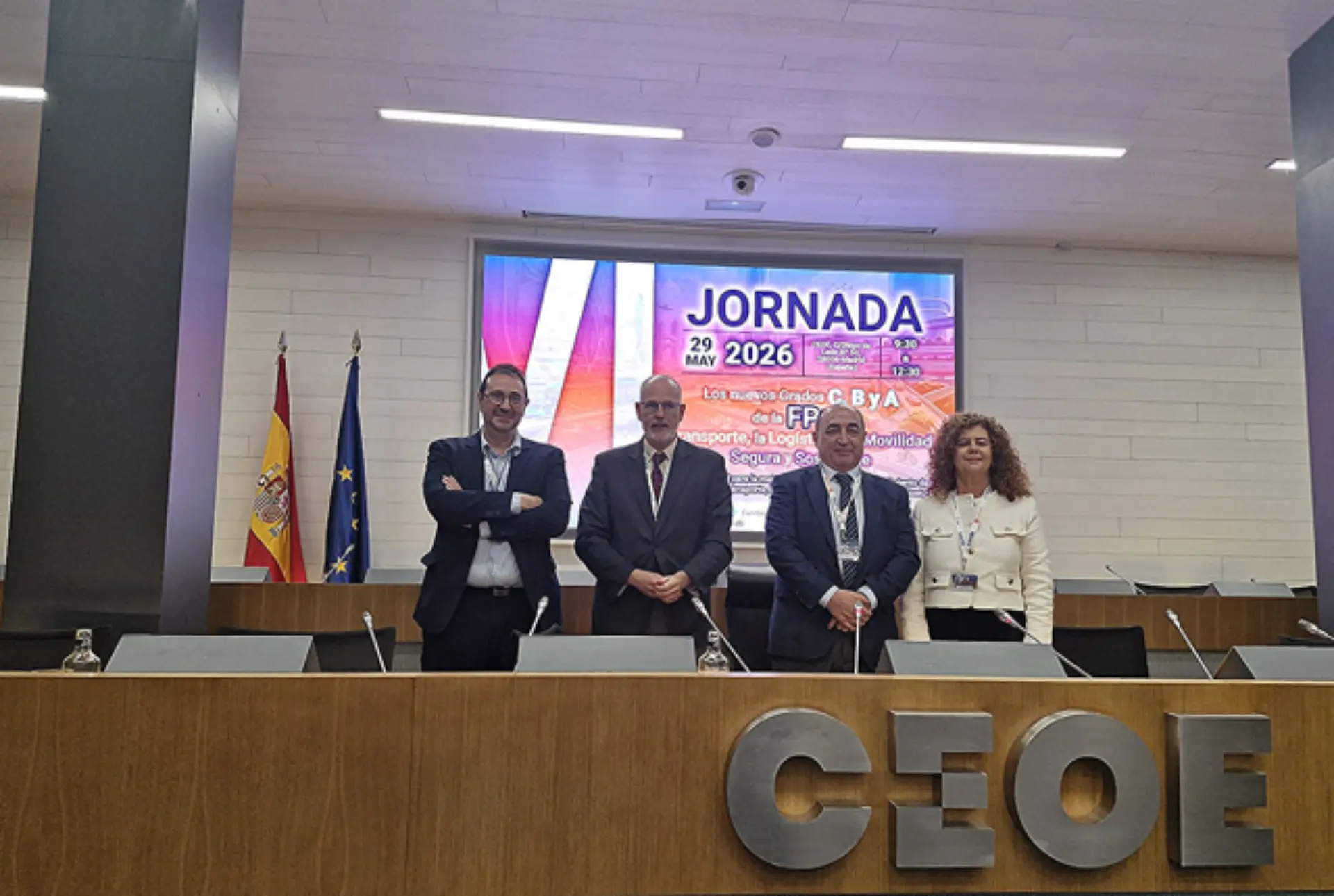 Cuatro personas están de pie detrás de un escritorio en una sala de conferencias con "CEOE" y una pantalla de presentación detrás de ellos, hablando de FP del transporte y de las oportunidades de formación en línea de la PAC.