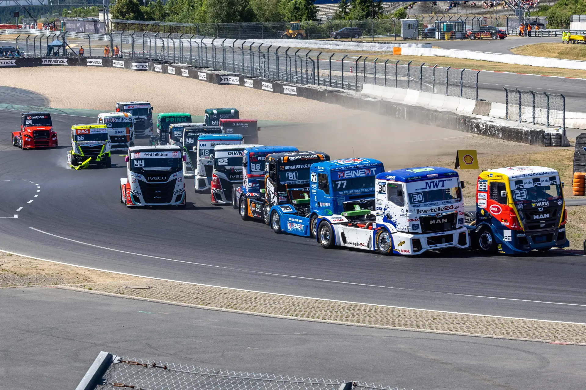 Varios camiones de carreras circulan a toda velocidad por una curva del circuito, con el rugido de sus motores de fondo, mientras compiten en una carrera de camiones «borrador automático» en un día soleado.
