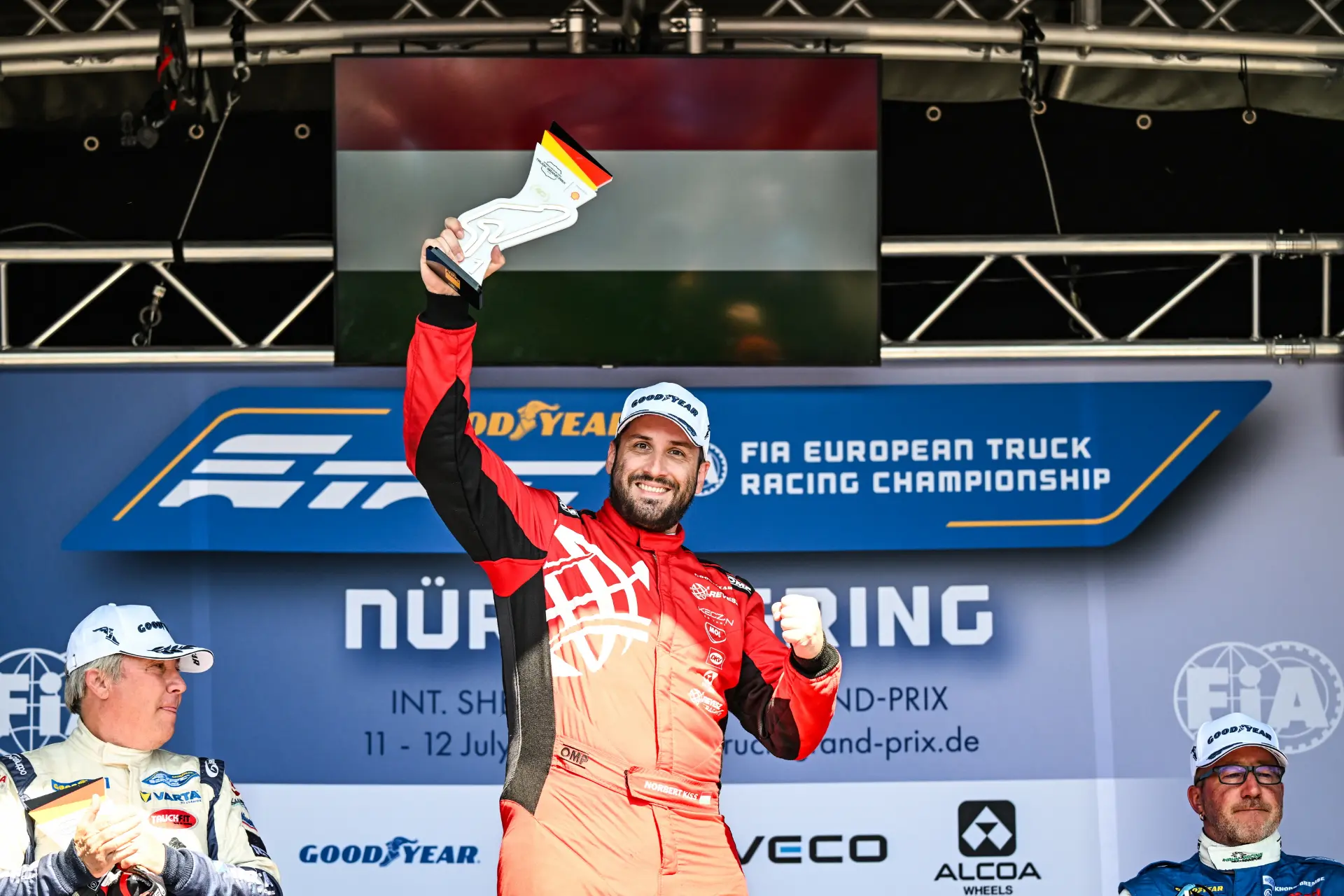 Un piloto de carreras, vestido con un mono rojo, levanta el trofeo en el podio, sonriendo, junto a sus rivales en una prueba celebrada en Nürburgring, mientras la cámara automática capta el momento de la victoria.