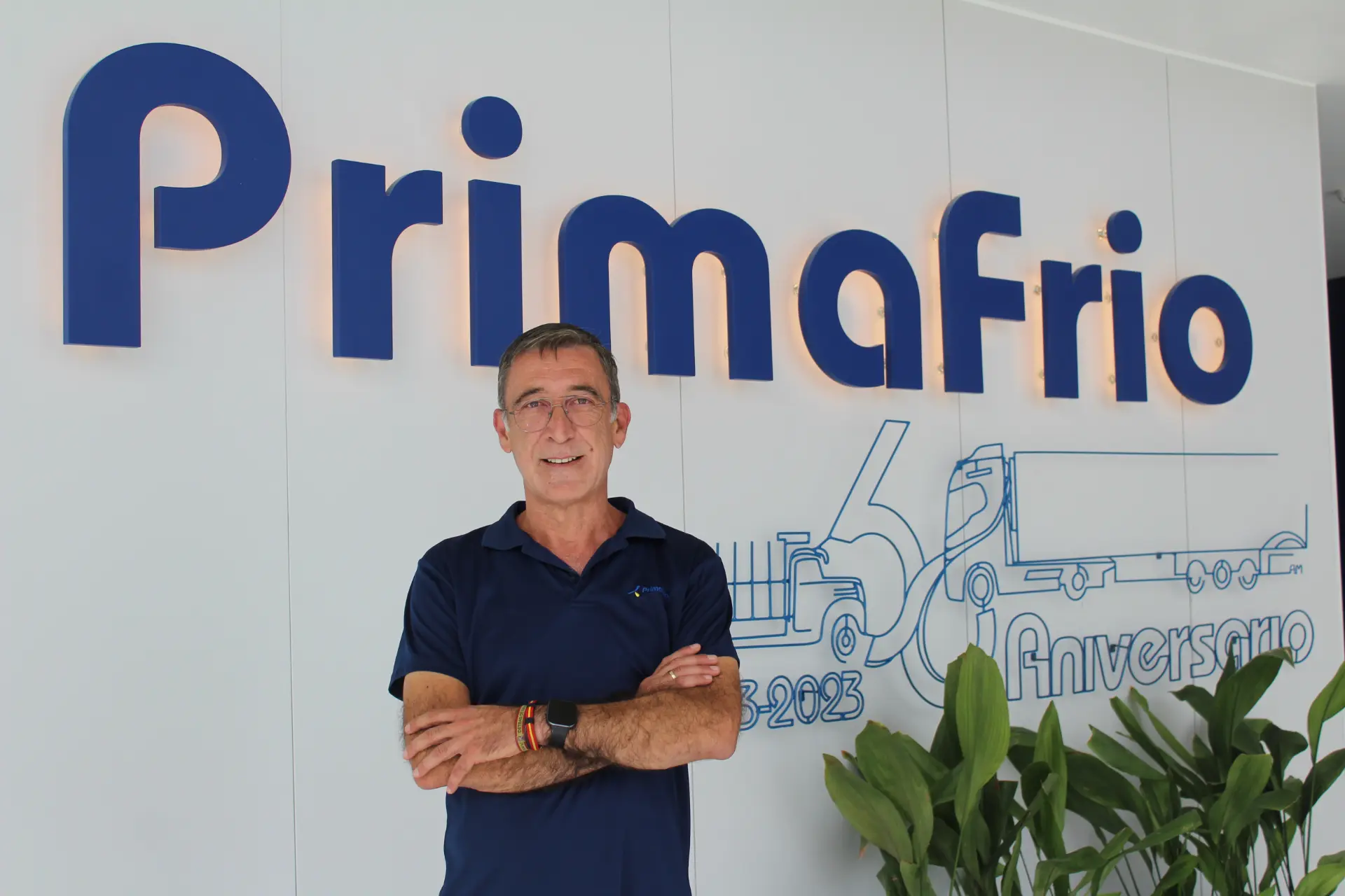 Un hombre de pie, con los brazos cruzados, frente a un gran logotipo de Primafrio y un mural conmemorativo del aniversario en una pared blanca, junto a un cartel visible que dice «Borrador automático».