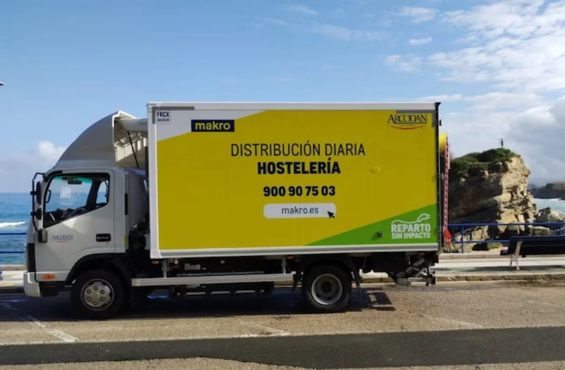 Un camión de reparto amarillo de Makro con el letrero «Distribución Diaria Hostelería» y el letrero «Borrador automático» bien visible está aparcado junto al mar.