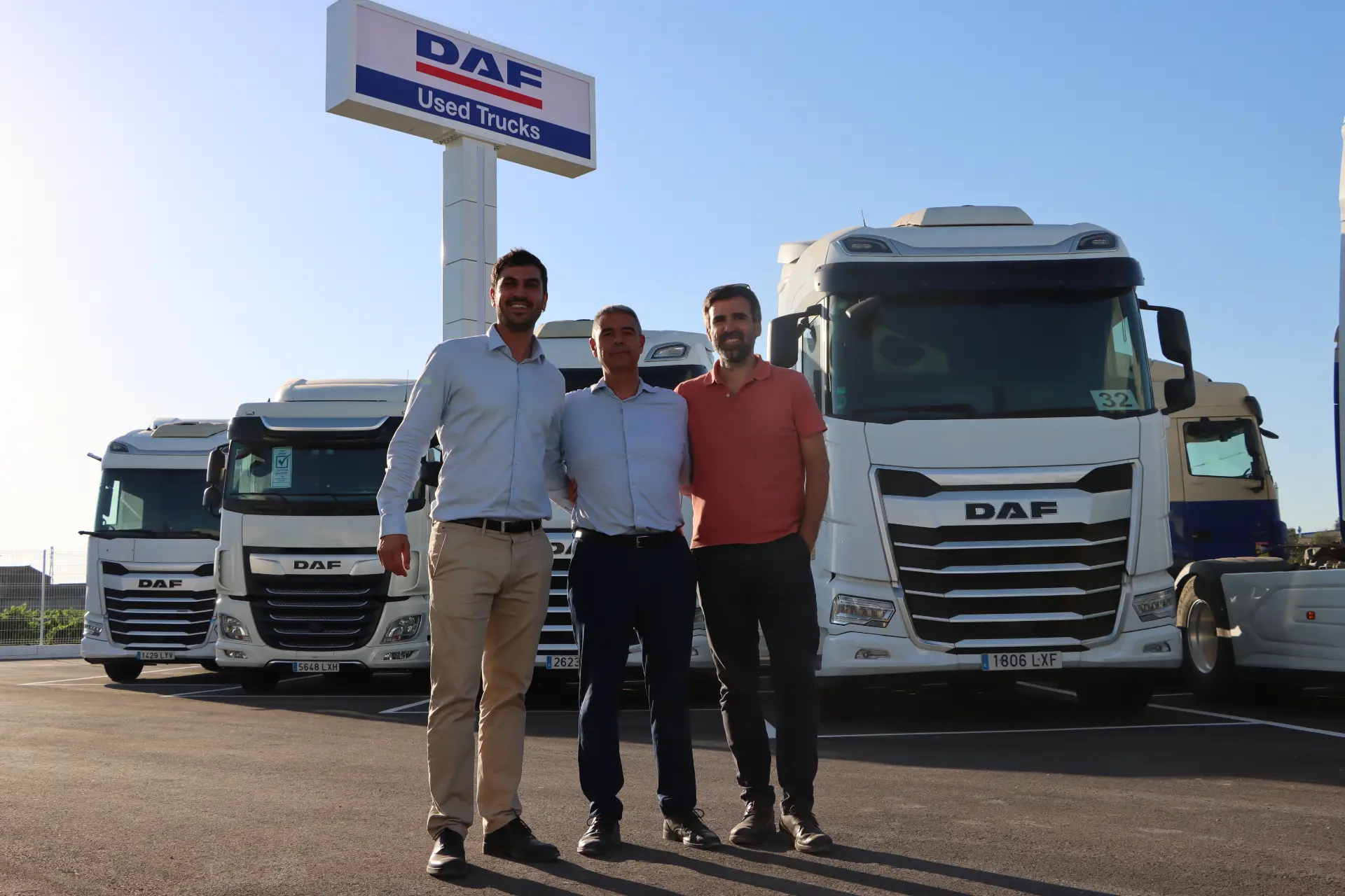 Tres hombres de pie delante de unos camiones DAF en el recinto de un concesionario, bajo un cartel que dice «Camiones DAF de segunda mano».