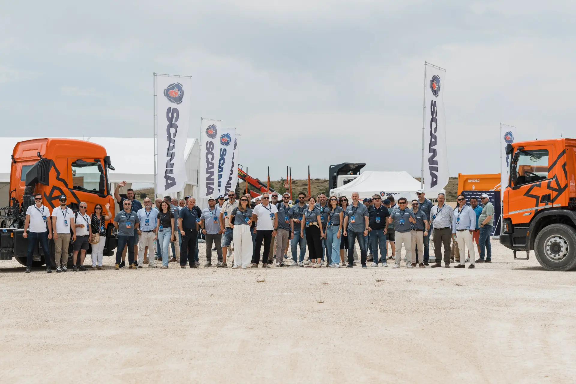 Un grupo de personas se encuentra al aire libre entre dos camiones Scania de color naranja, con banderas y carpas de Scania a sus espaldas, presentando «soluciones para obra» a clientes del sector de la construcción.