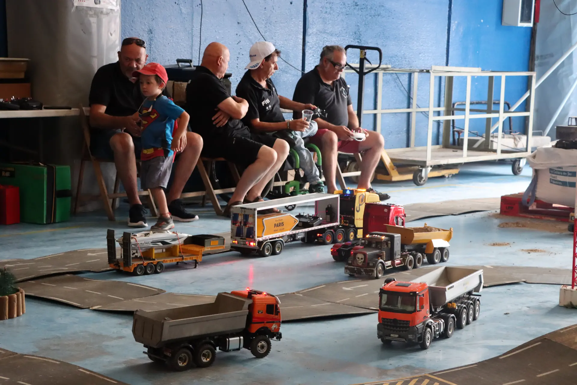 Unos adultos y un niño observan cómo circulan unos camiones teledirigidos por un circuito en miniatura montado en el interior, durante «La concentración de camiones de Olot», un animado encuentro celebrado en homenaje a Oti Cabadas.