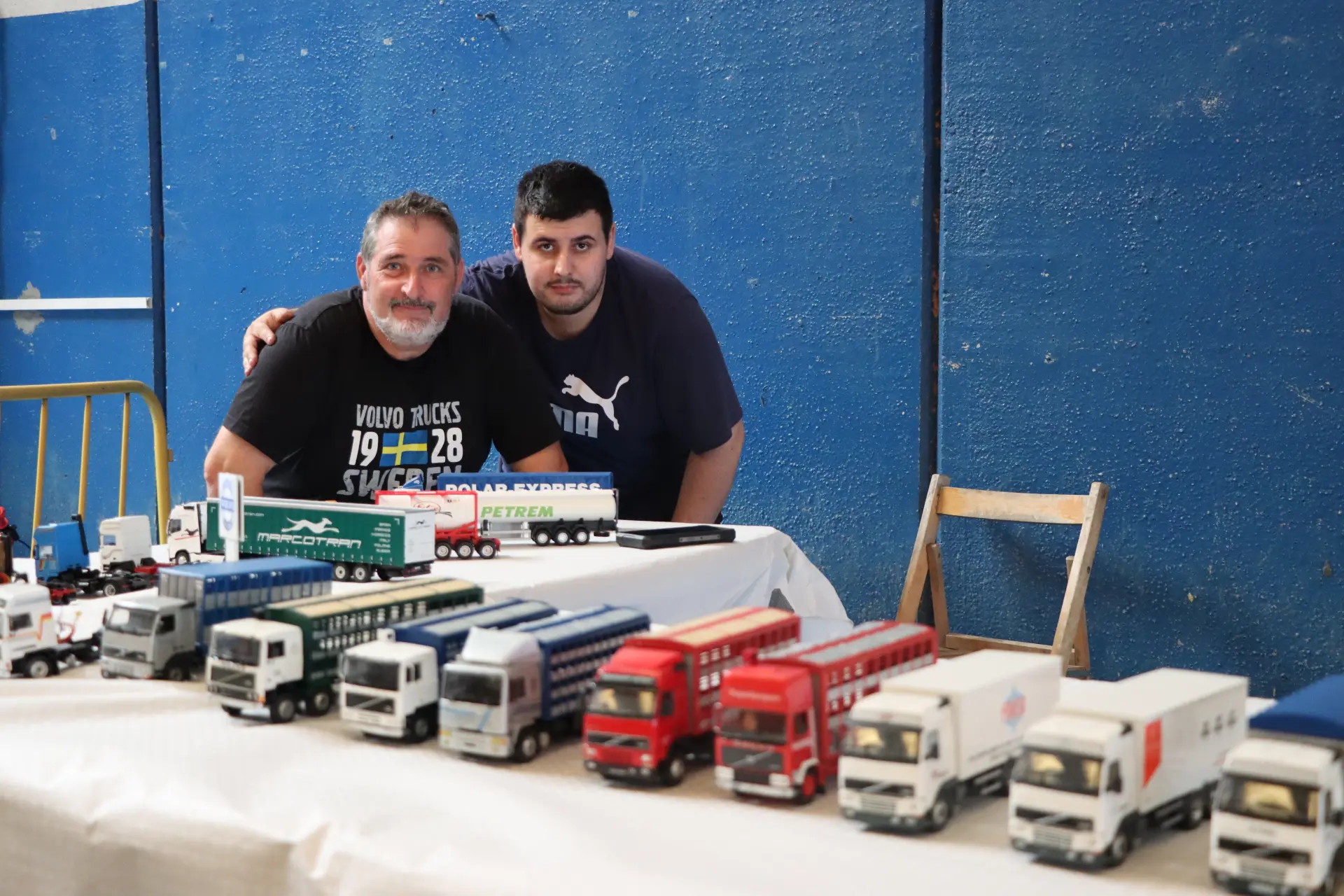 Dos hombres posan detrás de una mesa en la que se exponen maquetas de camiones, con una pared azul de fondo, en un evento celebrado en un recinto cerrado, en el que se rinde homenaje al equipo Truck Olot con un homenaje especial a Oti Cabadas.