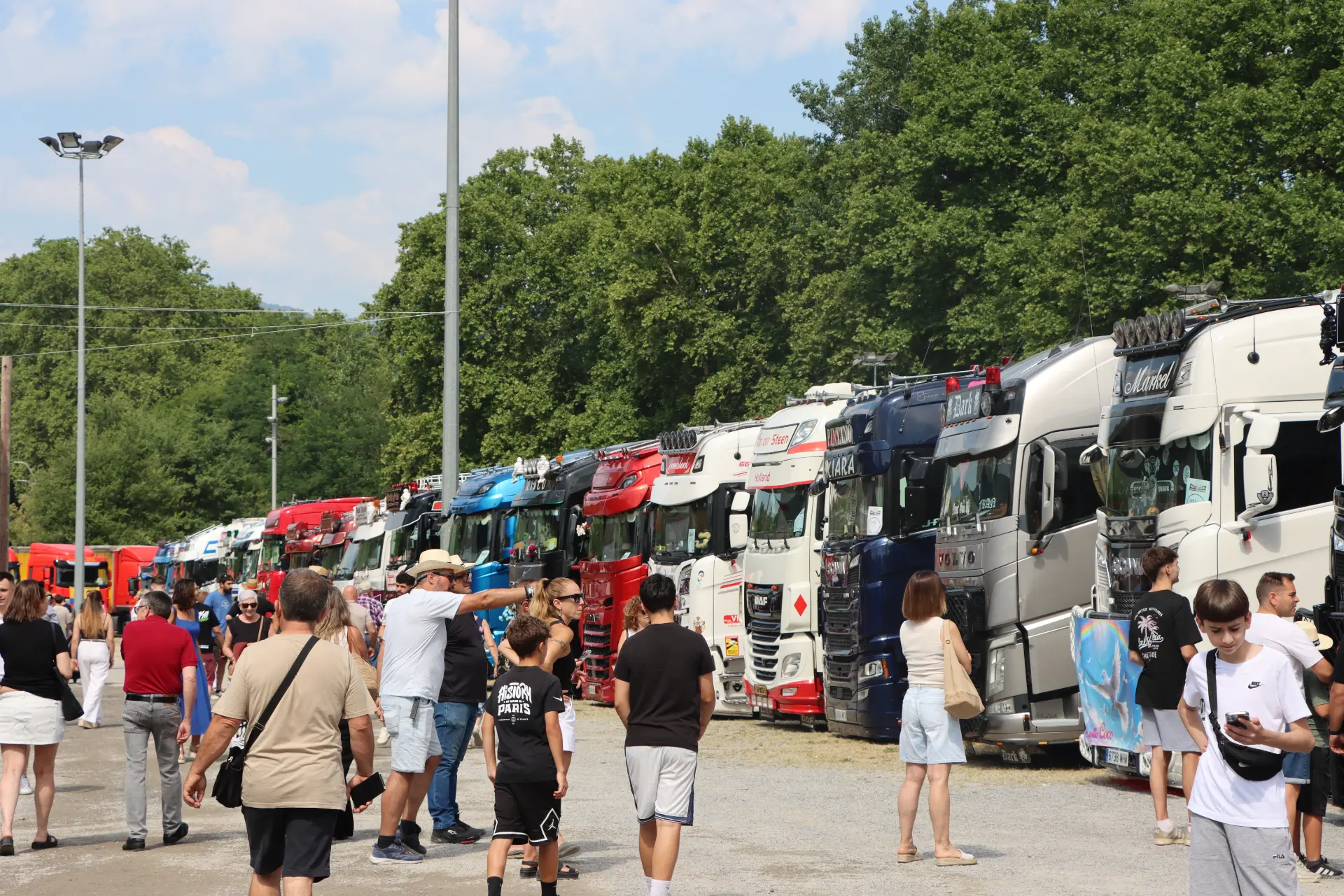 Gente que pasea junto a una fila de coloridos camiones aparcados en un evento al aire libre en un día soleado, disfrutando de la concentración de camiones de Olot organizada como homenaje a Oti Cabadas por Team Truck Olot.