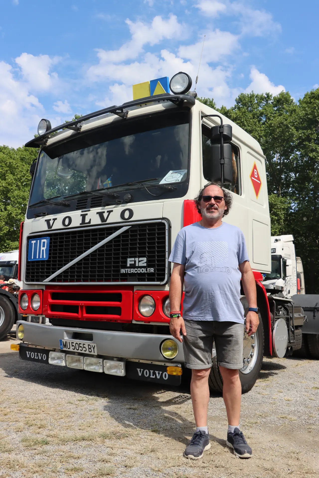 Un hombre en pantalones cortos posa sonriente delante de un camión Volvo F12 blanco en un día soleado en la concentración de camiones de San Cristóbal, sumándose al sincero homenaje a Oti Cabadas.