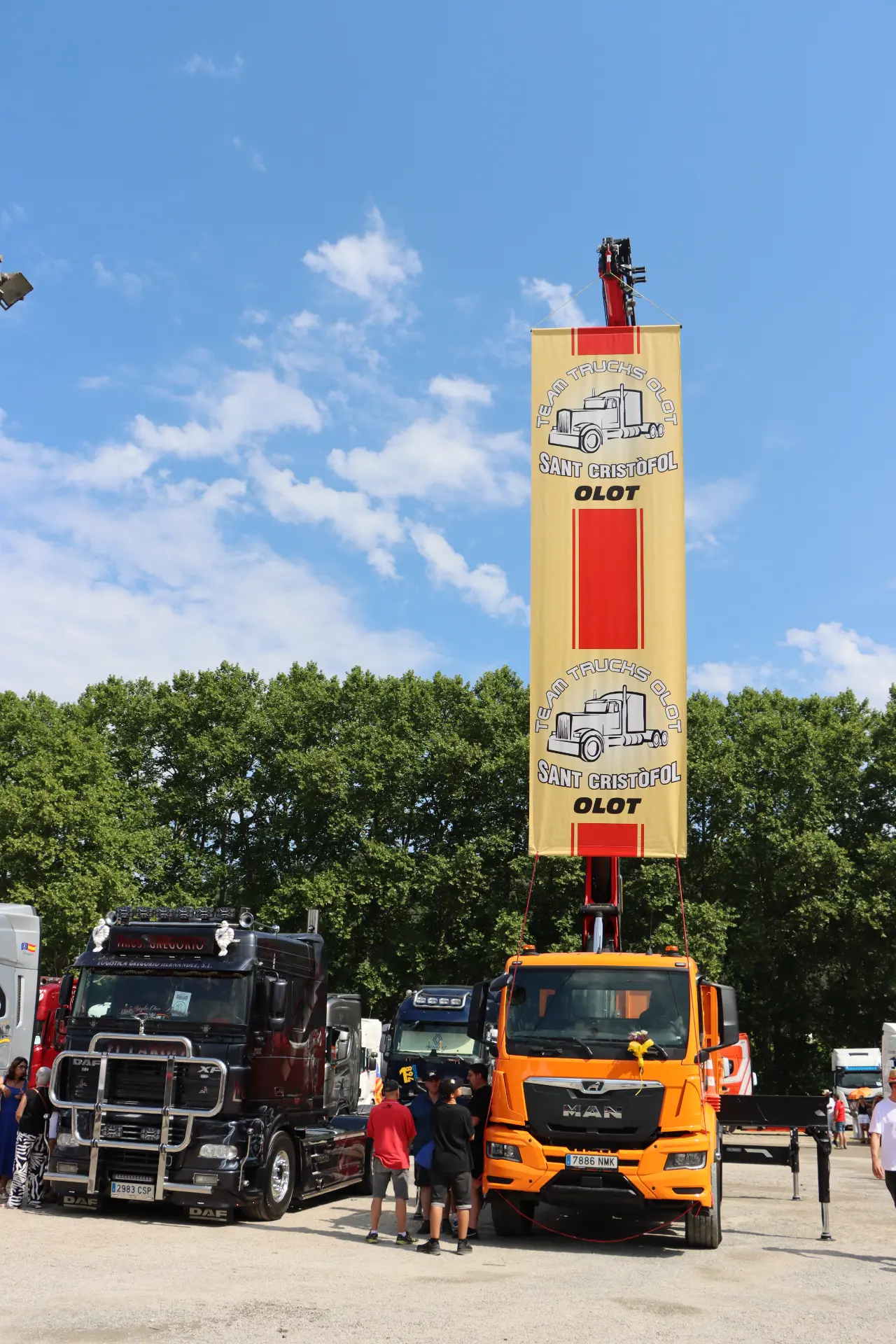 Dos camiones aparcados bajo una gran pancarta amarilla en la que se lee «Rally Trucks 4x4 Sant Cristofol OLOT» al aire libre, captando el espíritu del equipo de camiones de Olot en esta animada concentración.