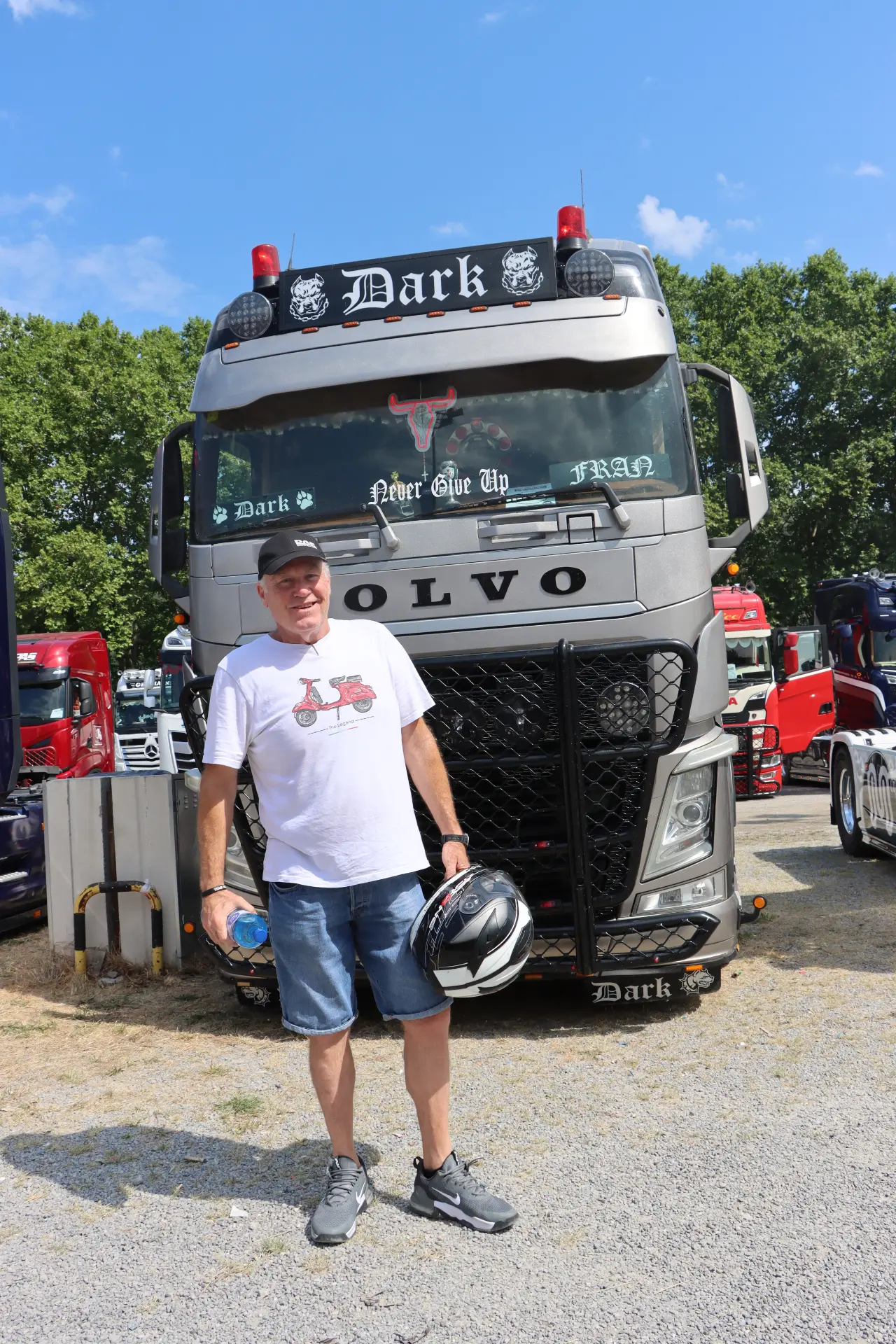 Un hombre sonriente, vestido con pantalones cortos, sostiene un casco y está de pie delante de un camión Volvo decorado en el evento «Concentración San Cristóbal Camiones».