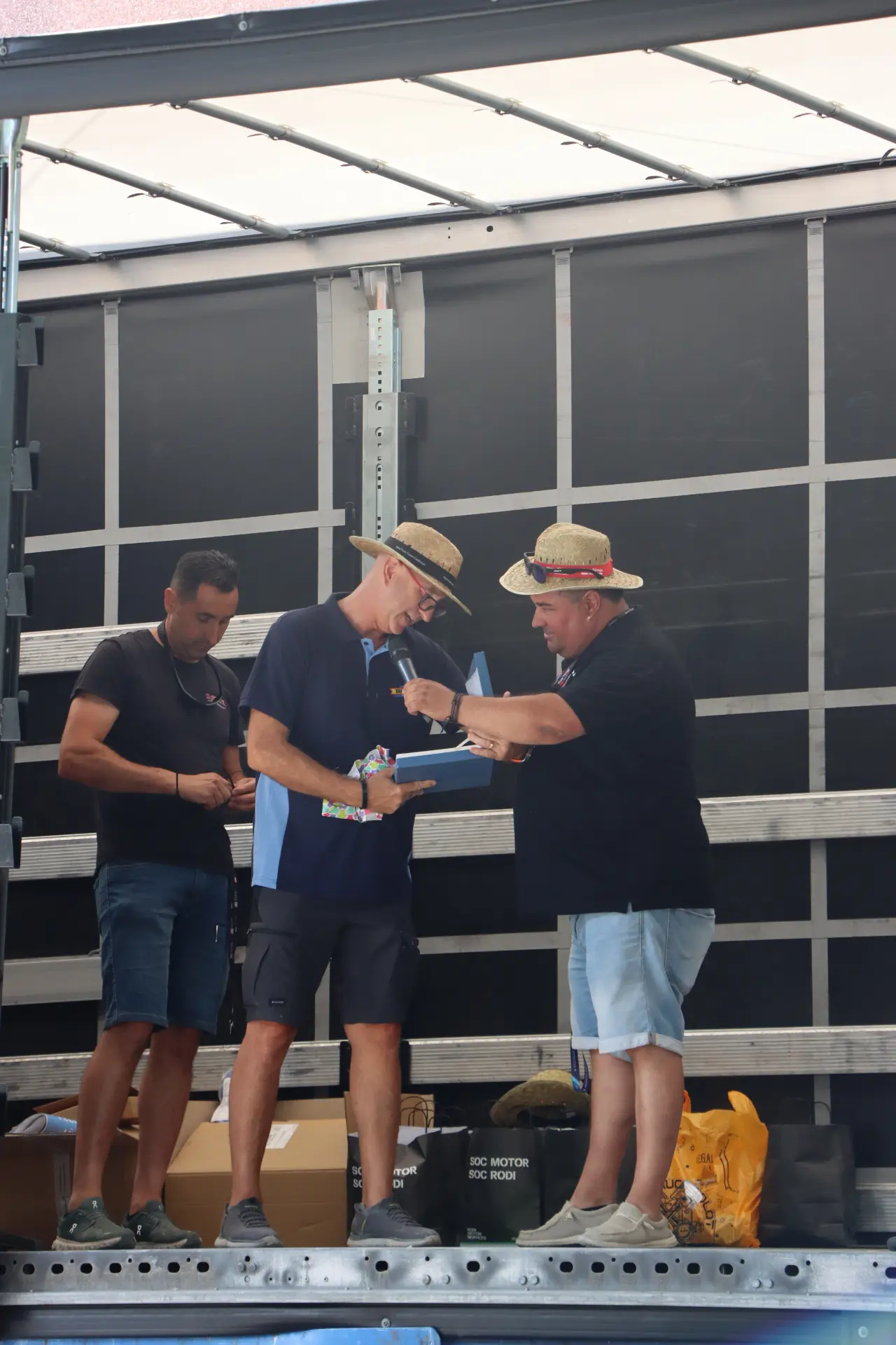 Tres hombres están de pie en la caja de un camión en la concentración de camiones de Olot: dos, con sombreros de paja, charlan, y uno, al fondo, mira su móvil; una escena muy apropiada durante el homenaje a Oti Cabadas.