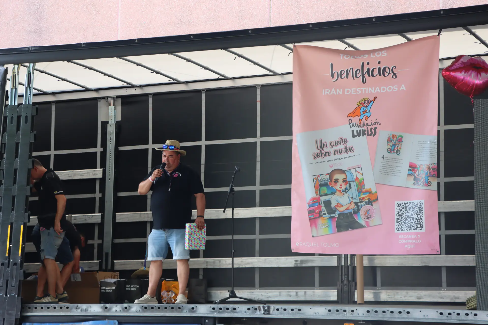 Un hombre con sombrero habla por un micrófono en el escenario, junto a una pancarta rosa de una organización benéfica y una pila de cajas, en la concentración de camiones de Olot.