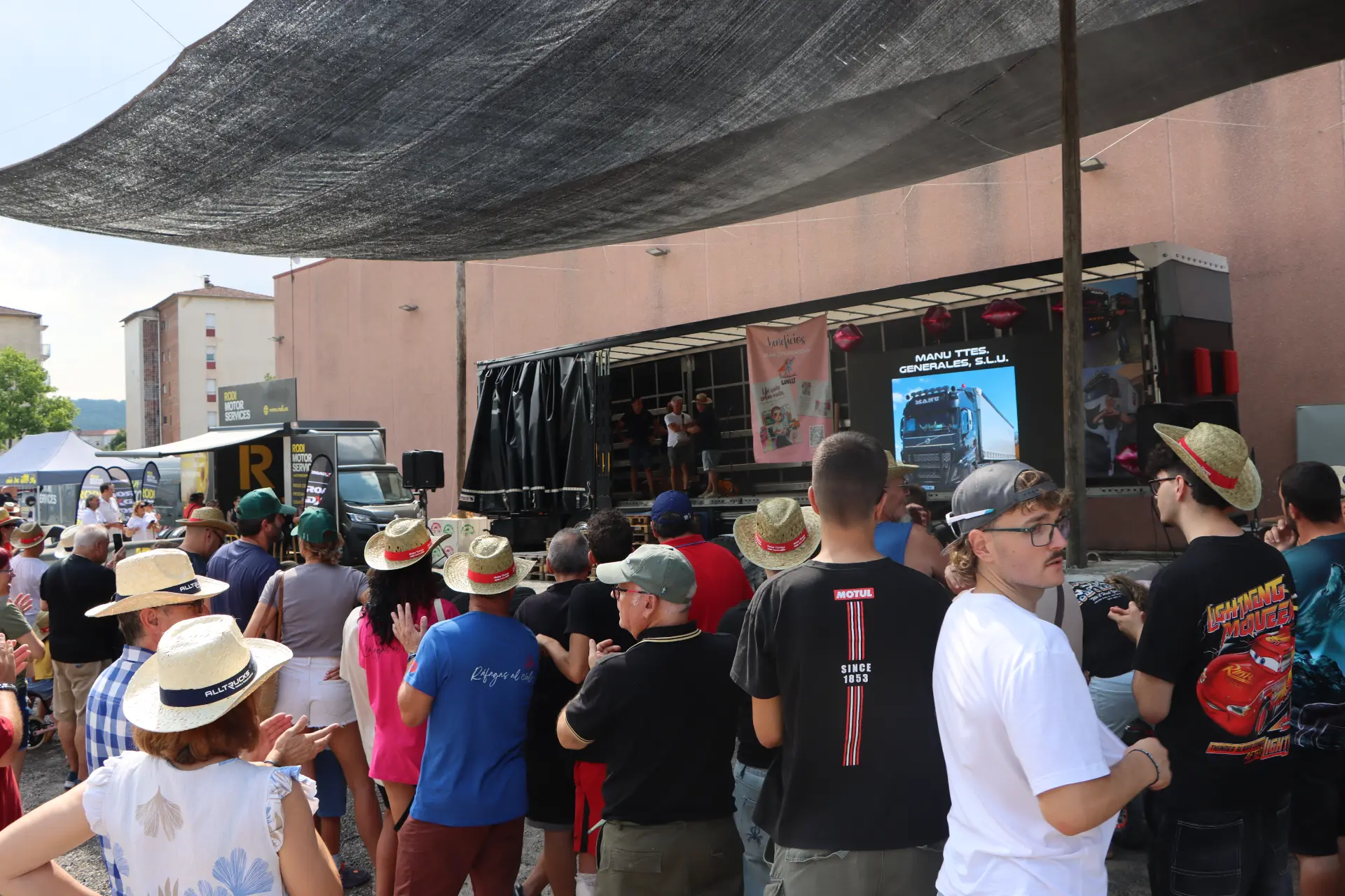 Un grupo de personas con gorras asiste a una actuación en un escenario al aire libre, bajo un gran toldo negro, en la «concentración de camiones de Olot», donde los miembros del equipo «Truck Olot» y «Cocotruckergirl» se reúnen para celebrar su pasión compartida por los camiones y la comunidad.