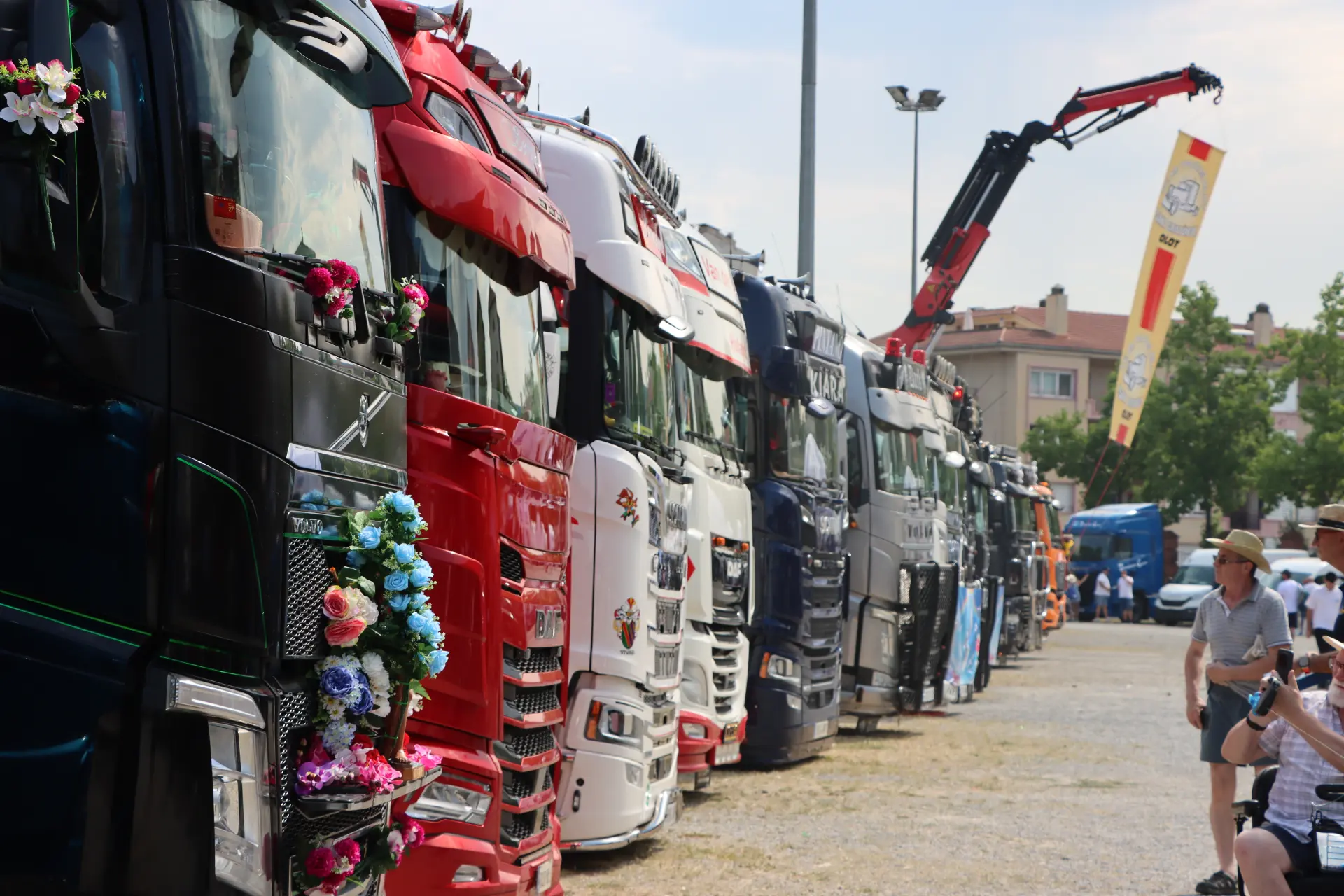 Una fila de camiones decorados con colores vivos, aparcados al aire libre durante una concentración, con gente paseando y sentada a su alrededor, constituye un animado homenaje a Oti Cabadas y al espíritu vibrante de «cocotruckergirl».