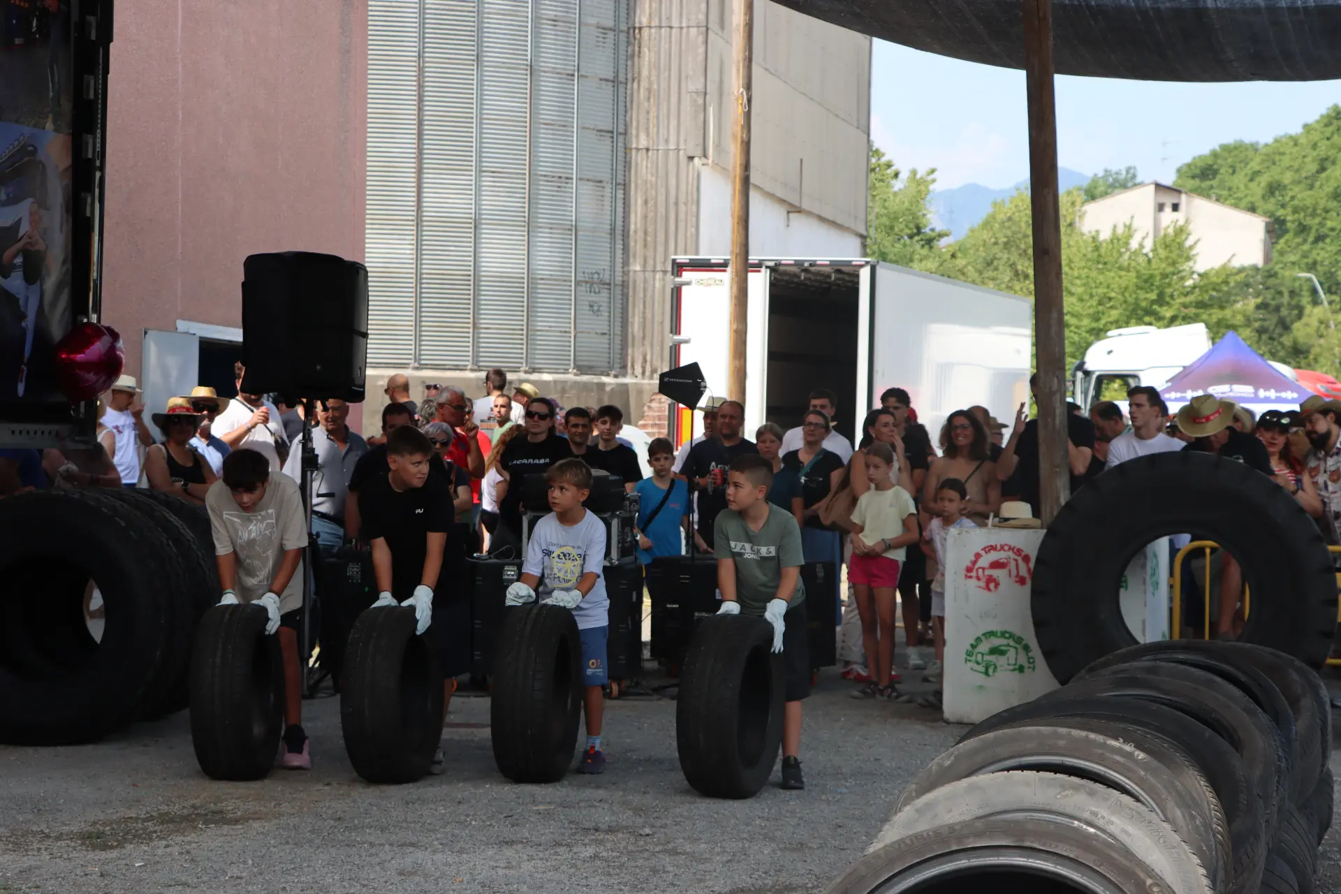 Cuatro niños hacen rodar neumáticos en una carrera ante una multitud que observa la escena en un evento al aire libre, que recuerda el animado ambiente que se respira en la concentración de camiones de Olot.