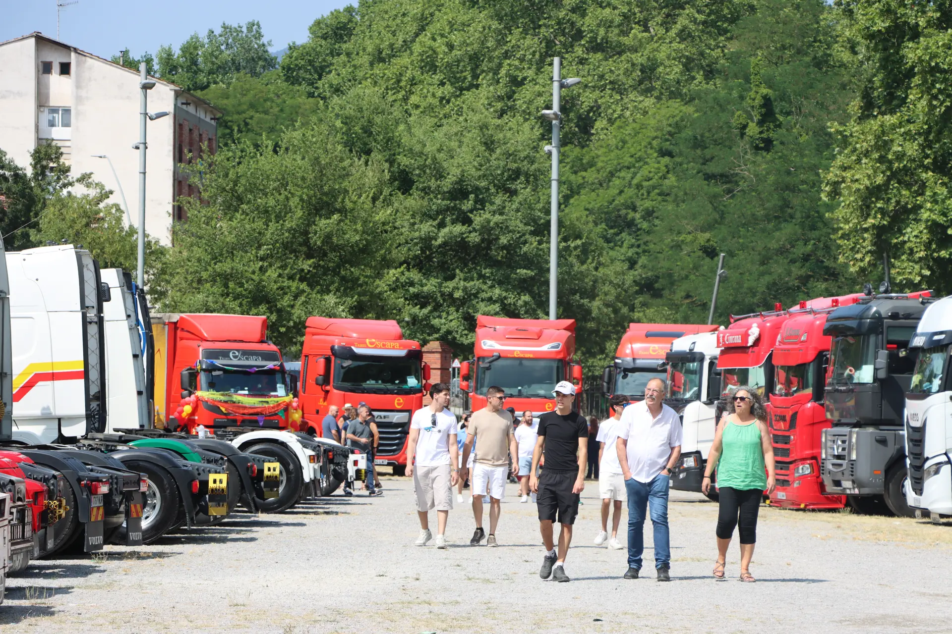 La gente pasea entre hileras de camiones rojos aparcados en un día soleado, con árboles y edificios al fondo, disfrutando del animado ambiente de la concentración de camiones de Olot.