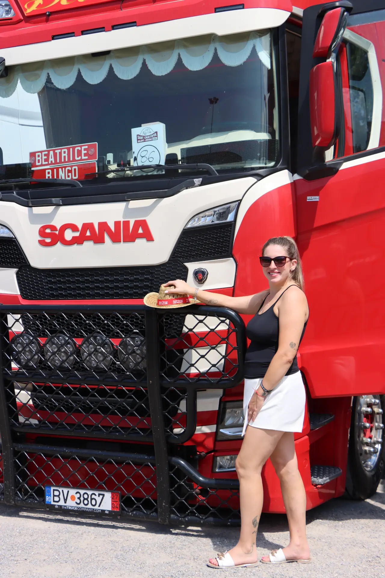 Una mujer con gafas de sol está de pie junto a un camión Scania rojo, con la mano apoyada en la parrilla delantera, durante la concentración con el equipo Truck Olot.