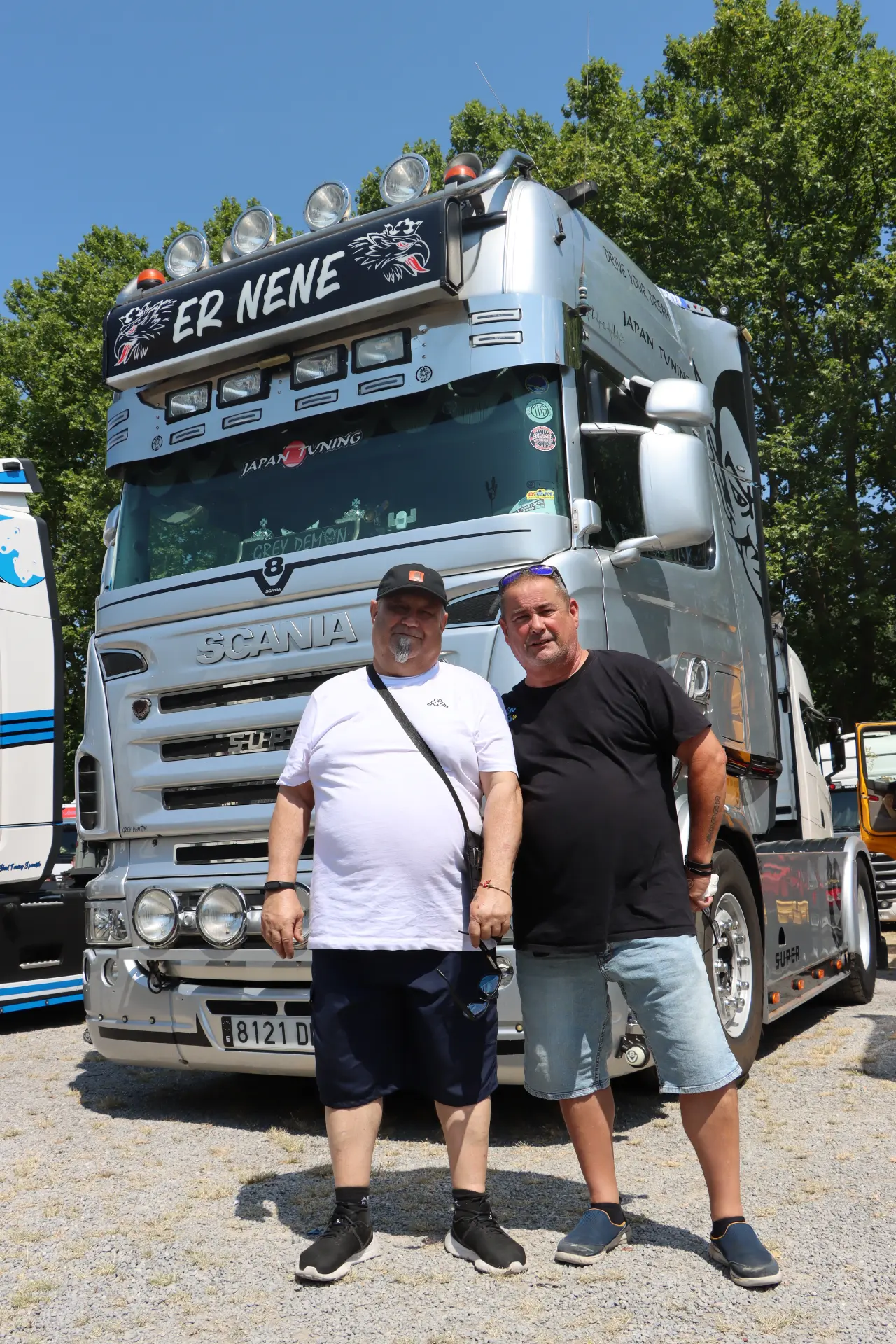 Dos hombres de pie frente a un gran camión Scania plateado en un día soleado, captando el espíritu de la concentración de camiones de San Cristóbal.