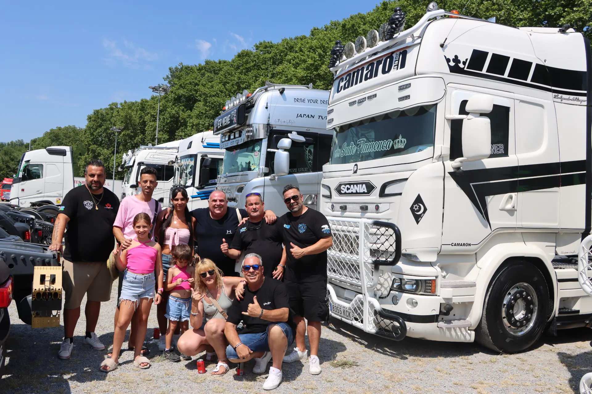 Un grupo de personas posa y sonríe delante de unos grandes camiones decorados en un día soleado durante la concentración de camiones de Olot, rindiendo homenaje a Oti Cabadas junto a cocotruckergirl.
