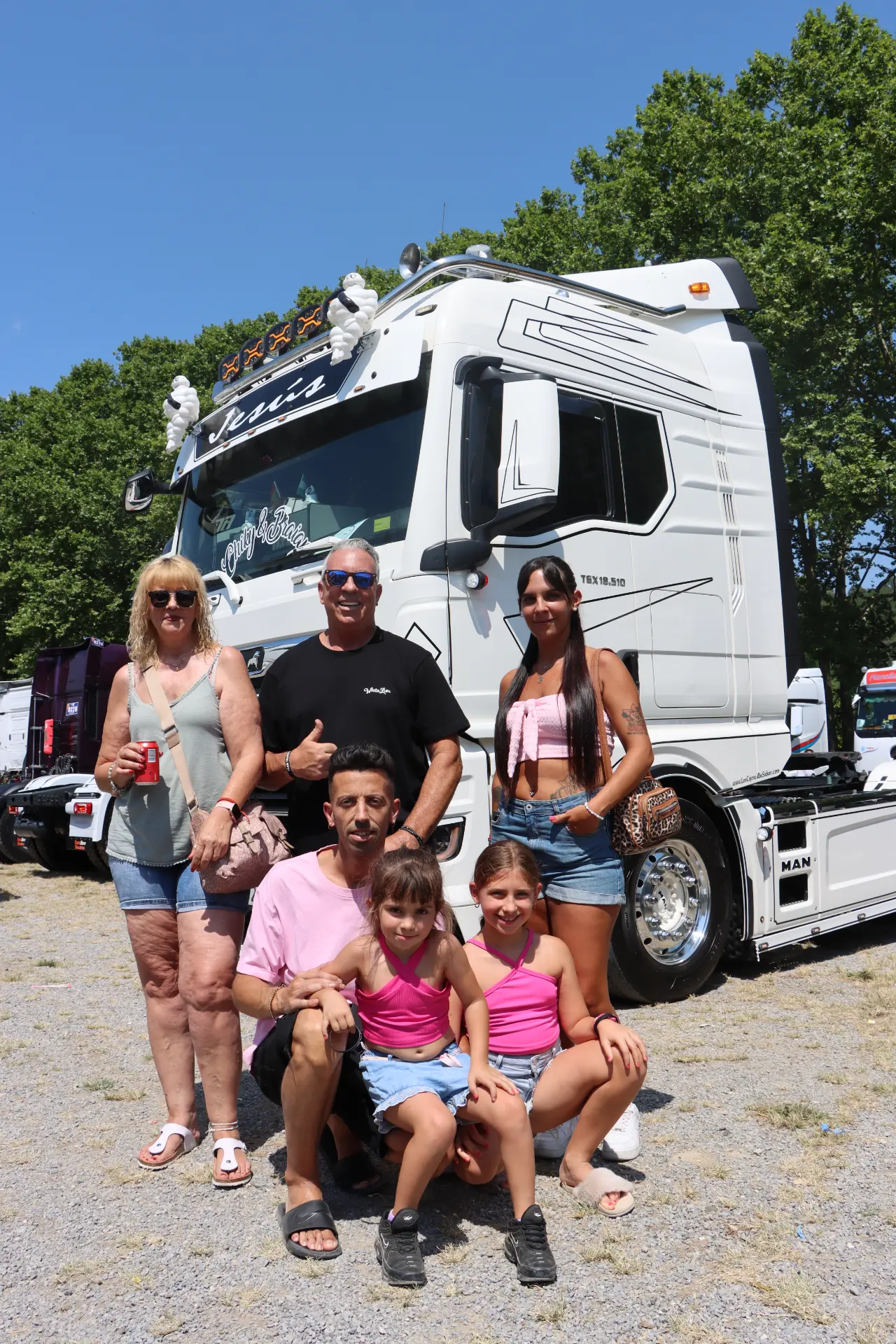 Una familia posa y sonríe delante de un gran camión blanco en un día soleado, captando el espíritu alegre de la concentración de camiones de Olot y rindiendo homenaje a Oti Cabadas con su entusiasmo por el evento.