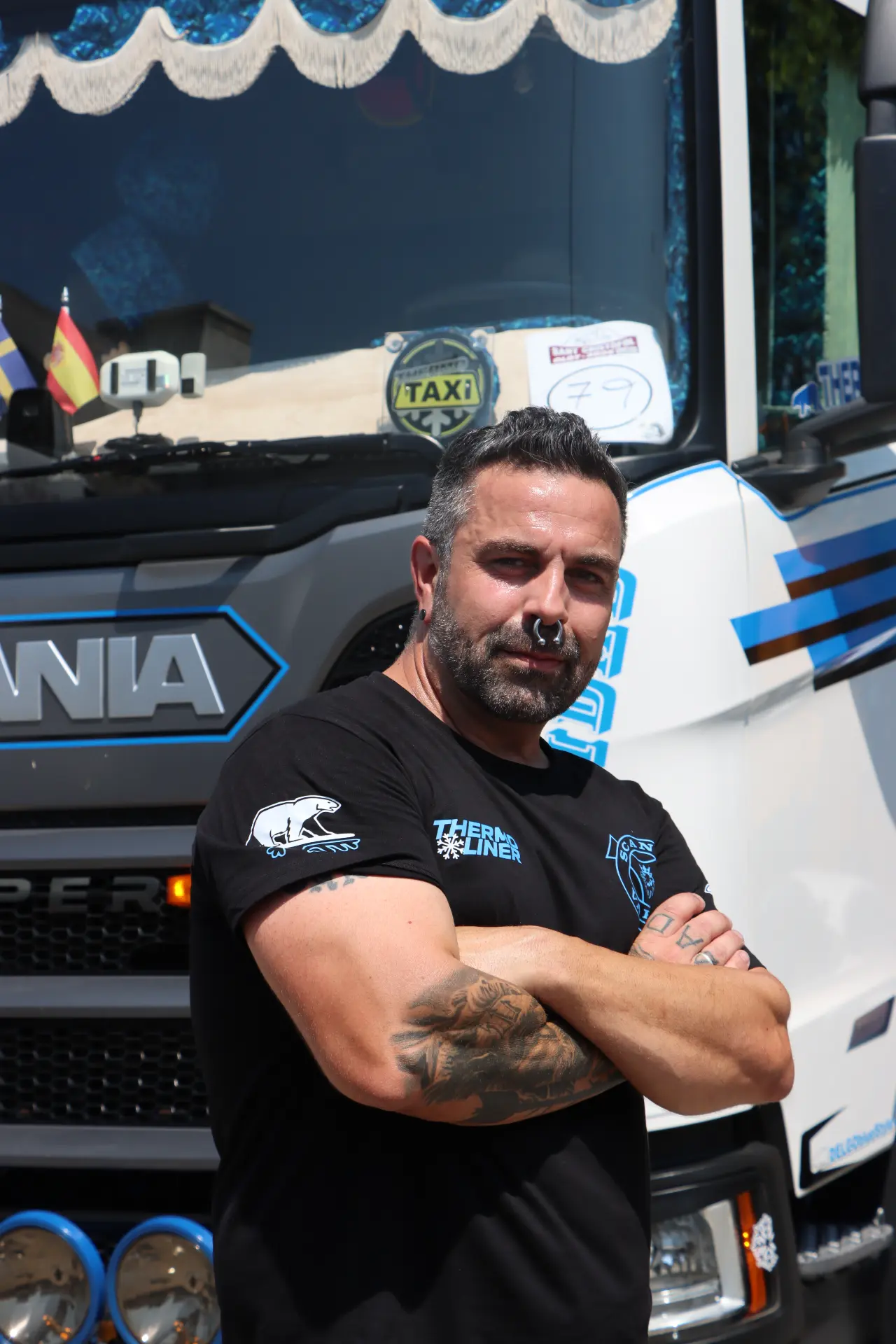 Un hombre con tatuajes y piercings posa con aire seguro frente a un camión Scania, con los brazos cruzados, en una imagen captada durante la concentración de camiones de Olot, un encuentro que también sirvió como homenaje a Oti Cabadas.