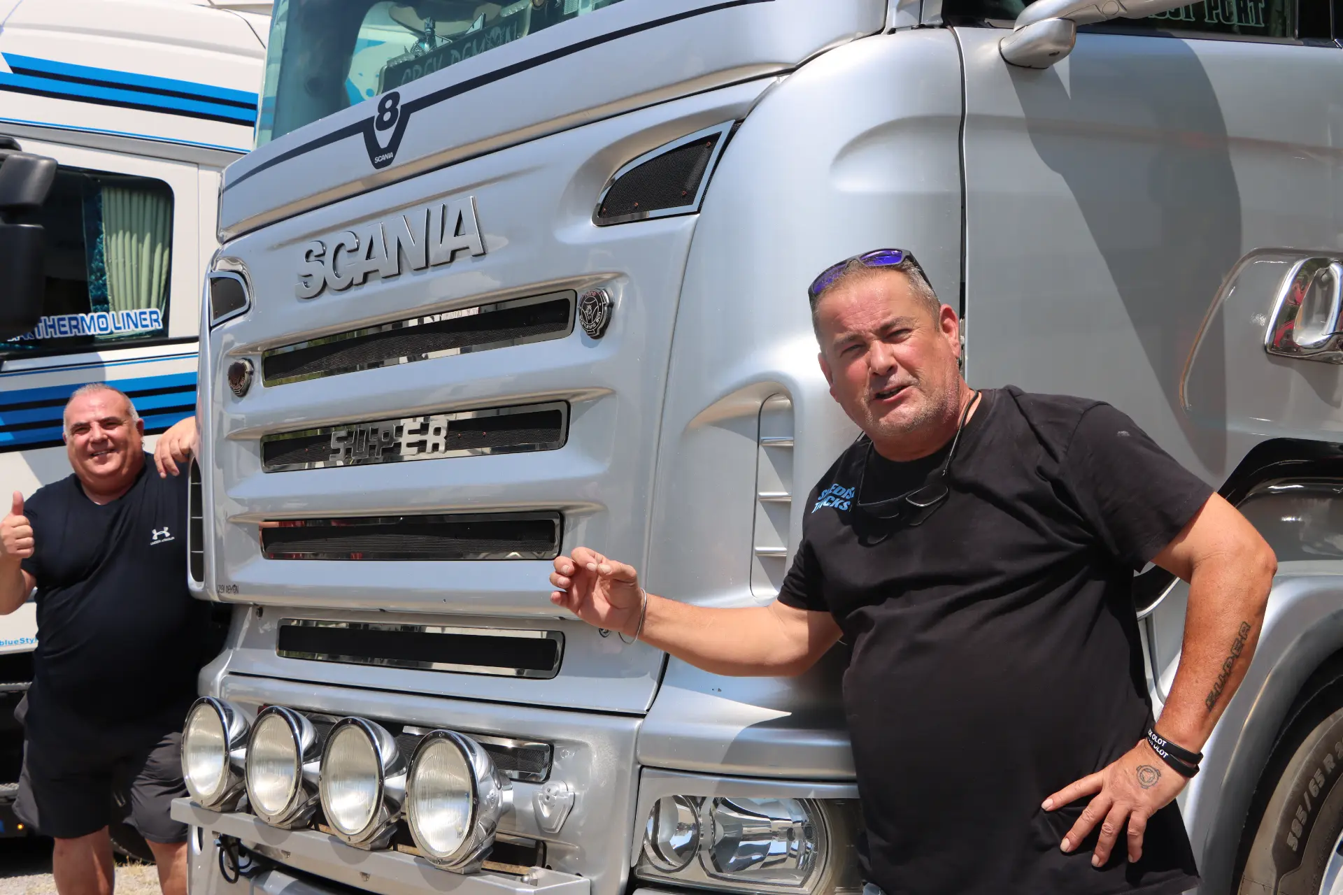 Dos hombres con camisetas negras posan junto a un gran camión Scania plateado en un día soleado durante la concentración de camiones de Olot.