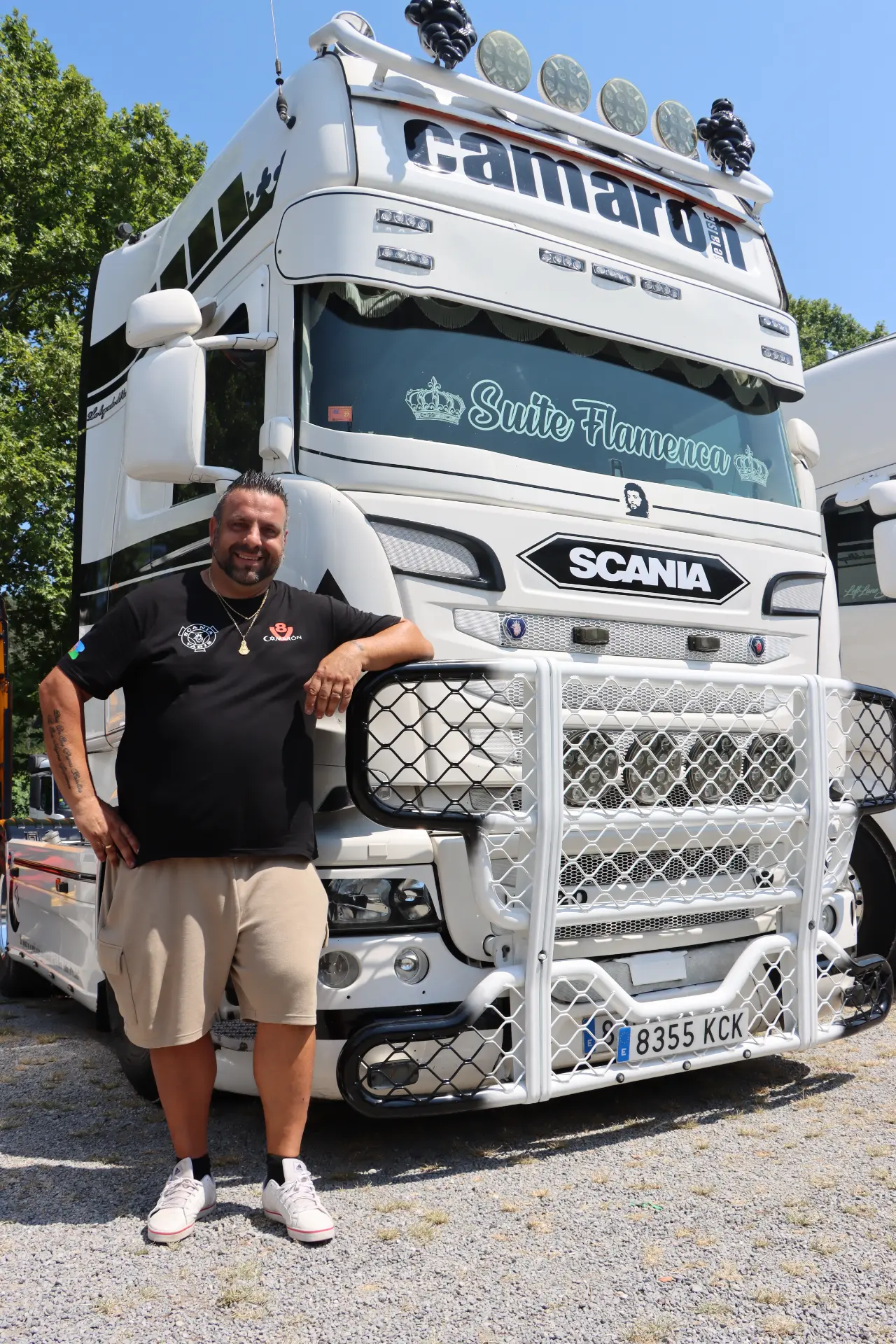 Un hombre sonriente, vestido con pantalones cortos, posa junto a un camión Scania blanco decorado, con protectores en la parrilla y letreros personalizados, en una concentración de camiones en San Cristóbal.
